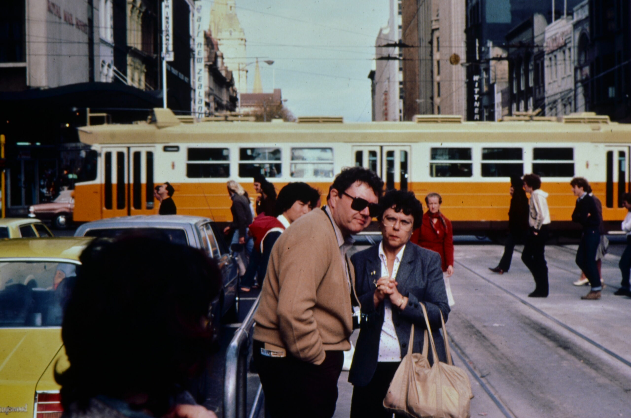 Busy urban street scene featuring a yellow-orange trolley bus across the road, with pedestrians in the foreground and tall buildings lining the street behind.