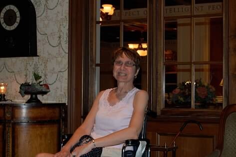 Older woman in a white sleeveless top sits in a wheelchair indoors, smiling.