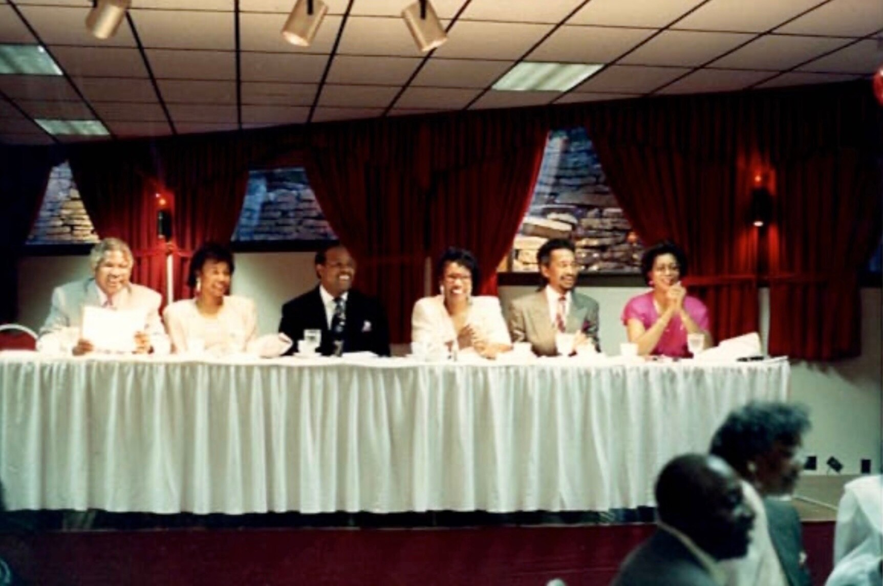 Panel of six people seated at a long, white-clothed table on a stage with red curtains behind, microphones and drinks in front of them.