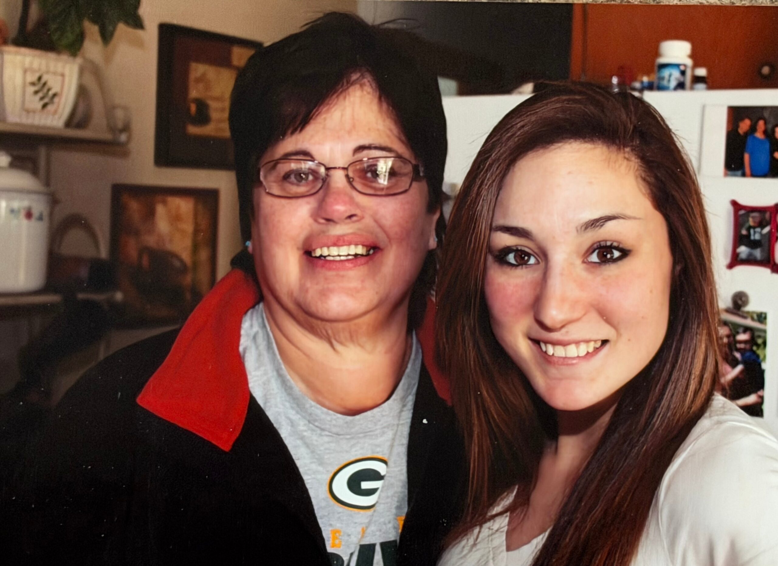 Two women smiling for a selfie in a home kitchen, one with short dark hair and glasses wearing a red scarf, the other with long brown hair.