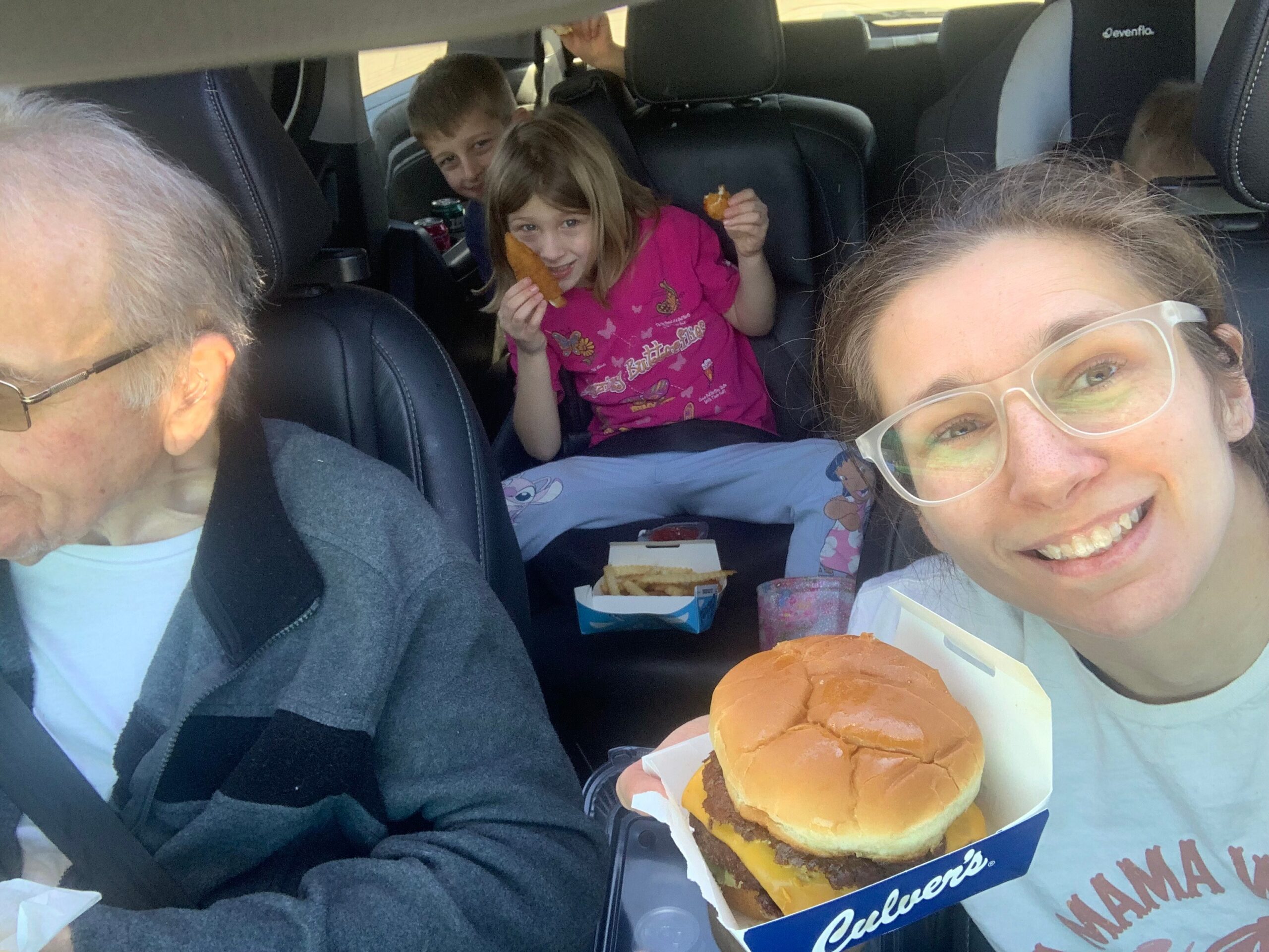Smiling woman with clear glasses taking a selfie in a car, with two kids in the back seat eating cheeseburgers and snacks nearby.