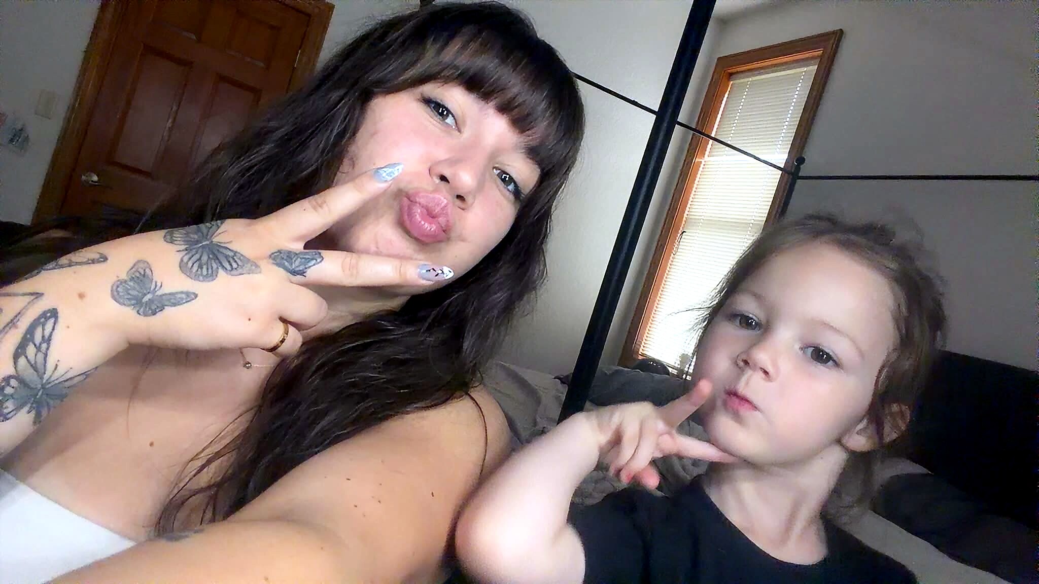 Two people pose for a selfie indoors: a woman with butterfly tattoos on her arm making a kissy face and a girl next to her pointing at the camera.