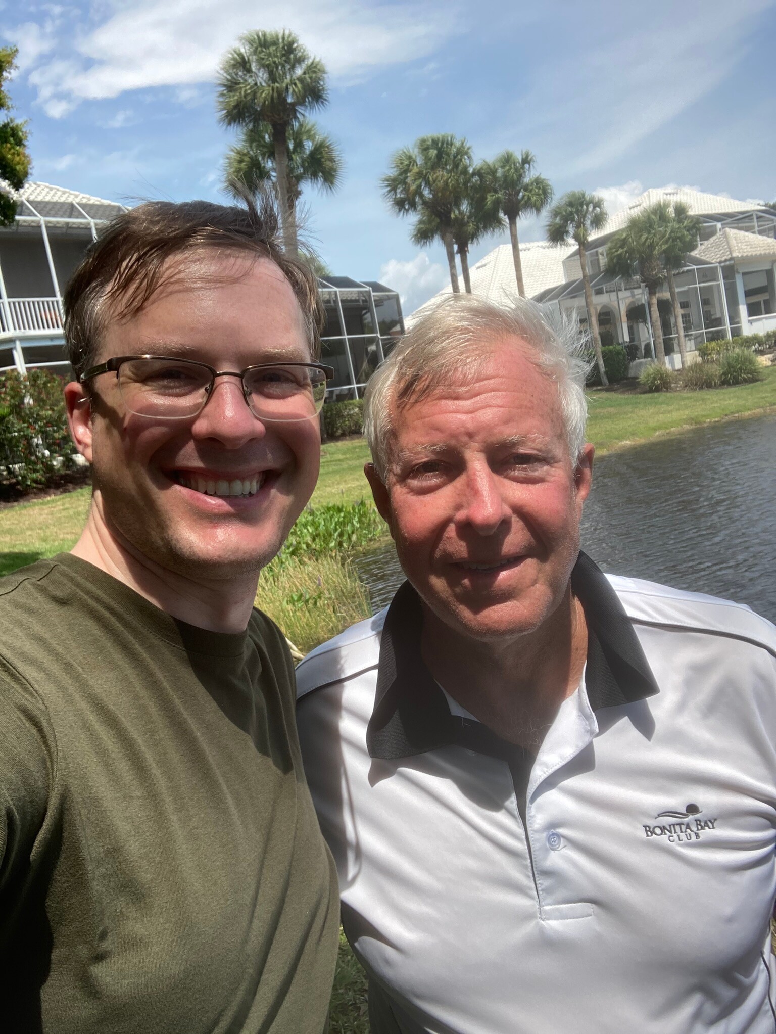 Two men smiling for a selfie by a canal with palm trees and white buildings in a sunny, resort-like area.