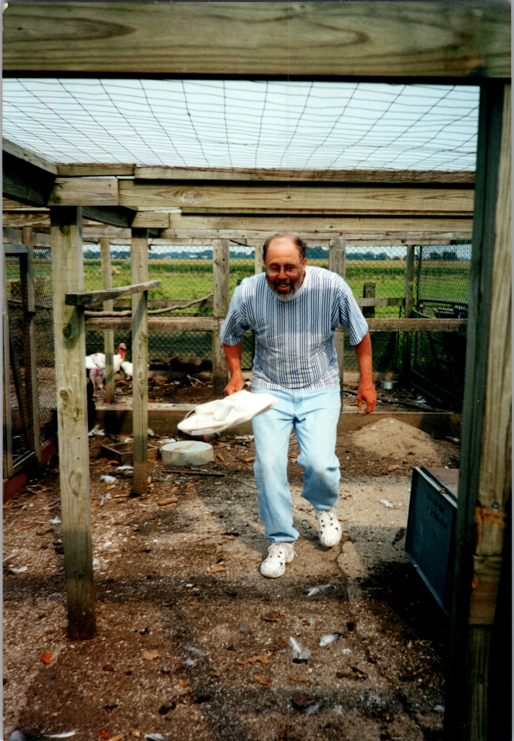 Older man in a striped shirt walks through a wooden chicken coop, holding a white cloth, outdoors on a farm.
