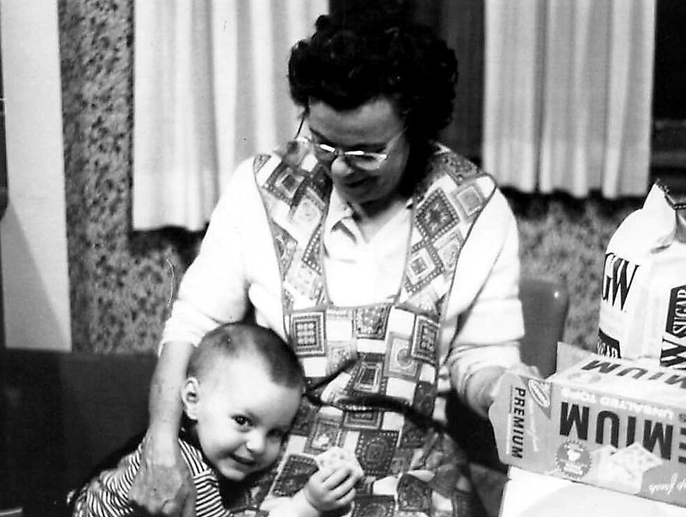 Grandmother wearing a patterned vest sits with a smiling baby on her lap indoors.