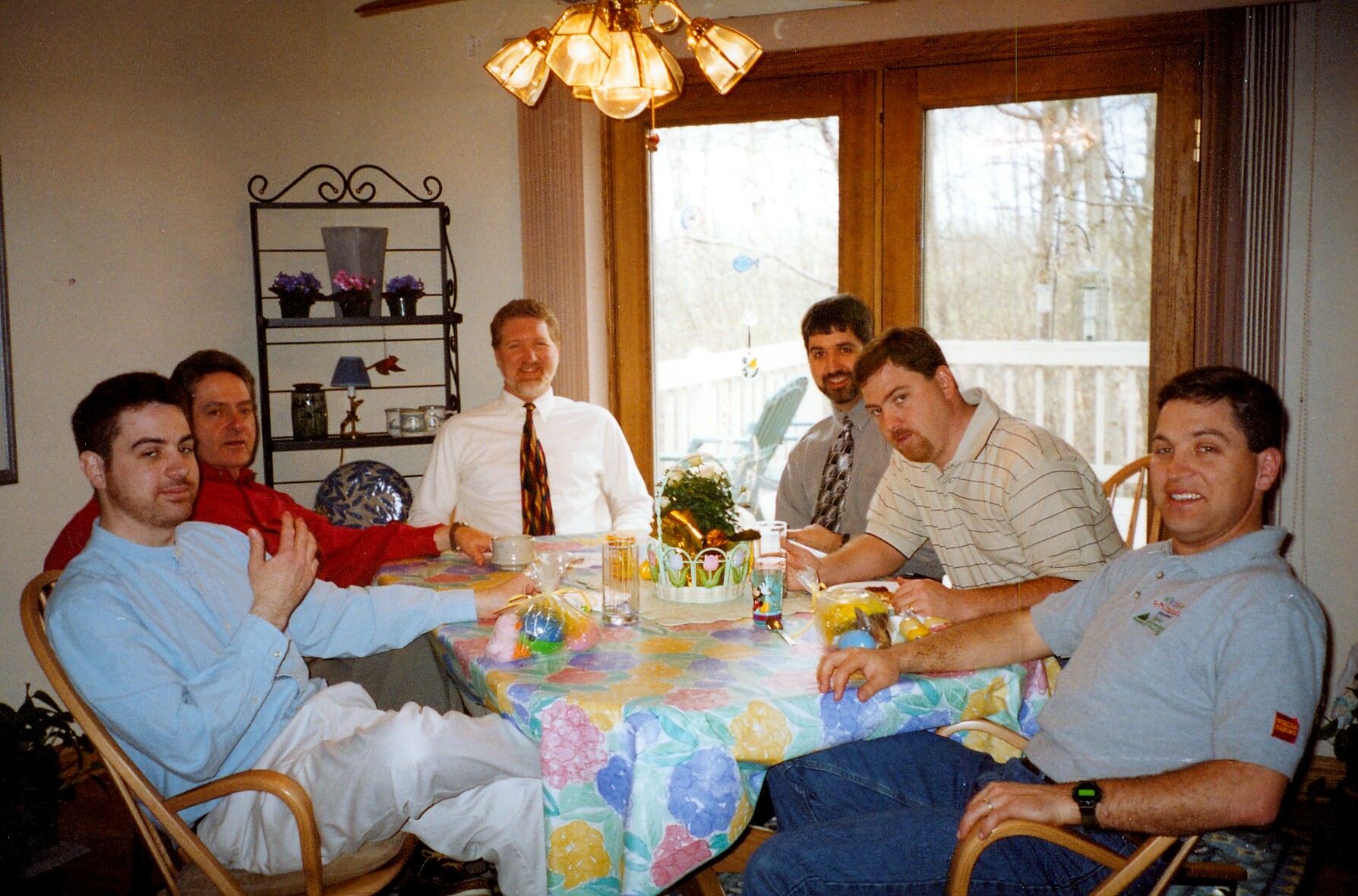 Six men sit around a table with a colorful tablecloth and Easter eggs, smiling at the camera in a dining room.