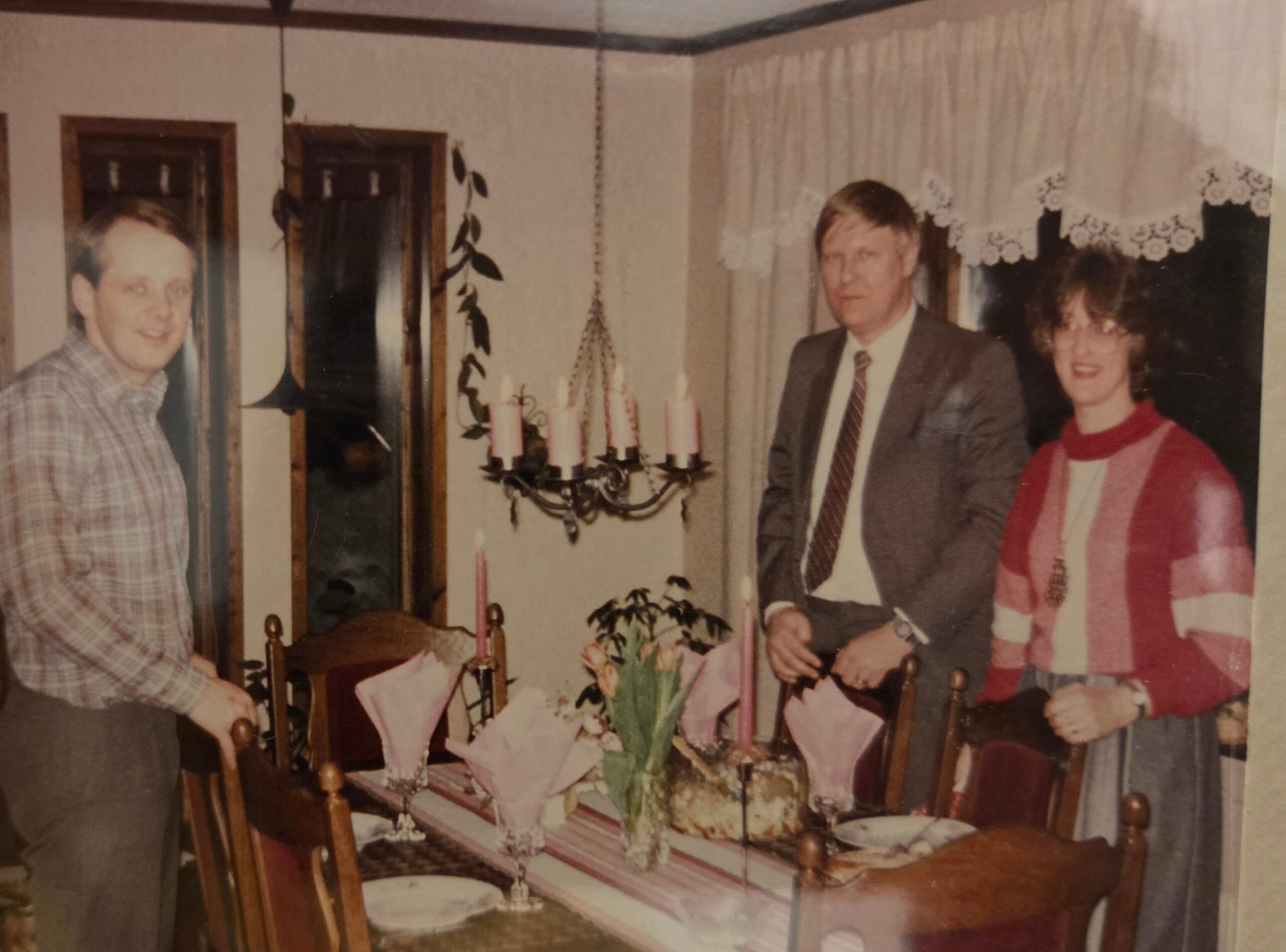 A man in a plaid shirt stands at a dining table with a birthday cake and candles, while two formally dressed people and a woman in red pose nearby inside a home.
