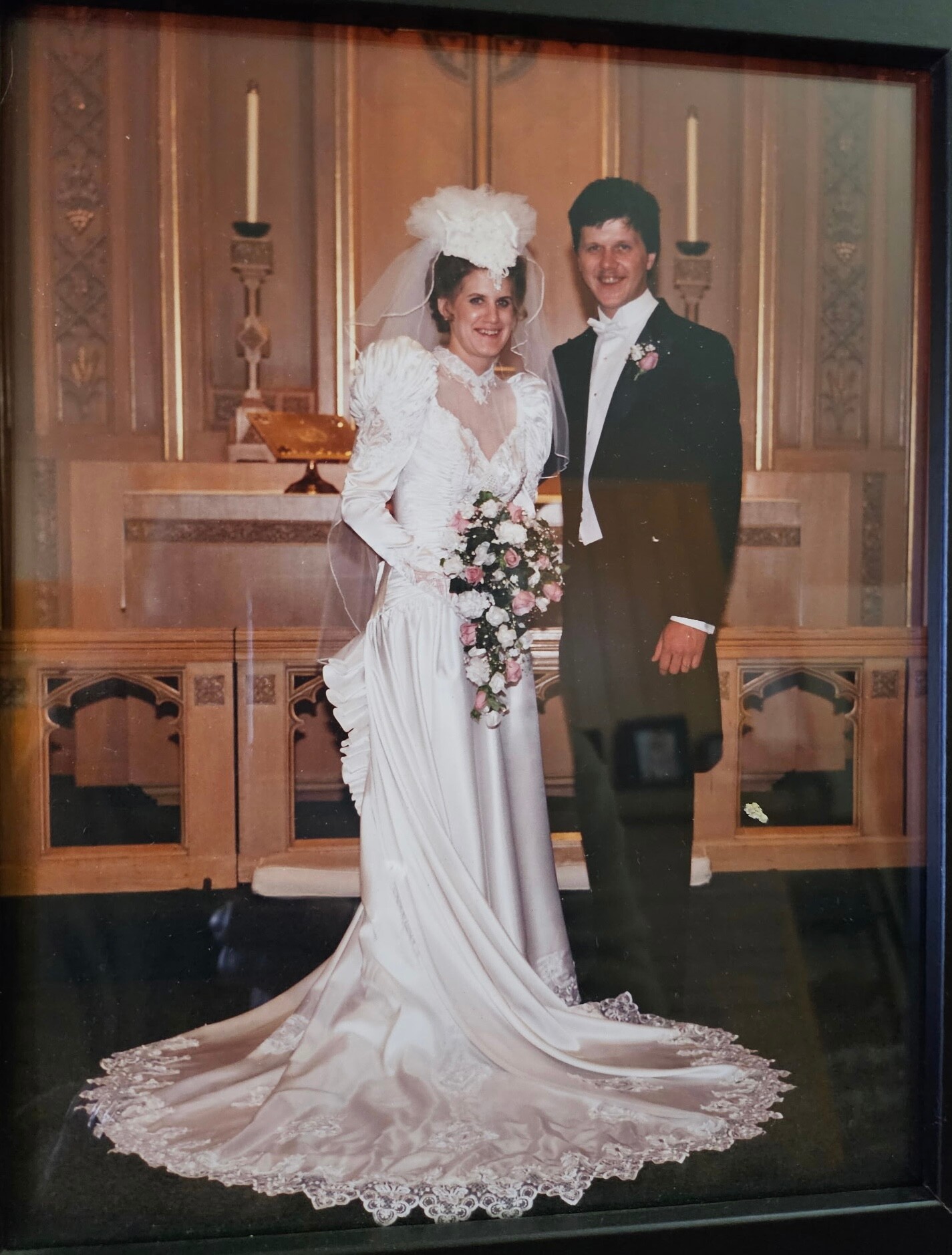 Bride in a white wedding gown with lace sleeves and a long lace-trimmed train, holding a pink-and-white bouquet, standing beside a man in a black tuxedo in a church setting.