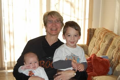 Older woman sits on a couch with a baby in her arms and a young boy beside her in a sunlit living room.