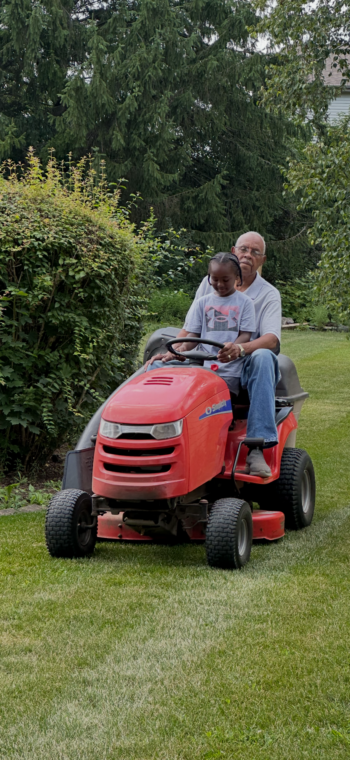 An elderly man and a young girl ride a red riding lawn mower together across a green backyard park-like yard with trees in the background.