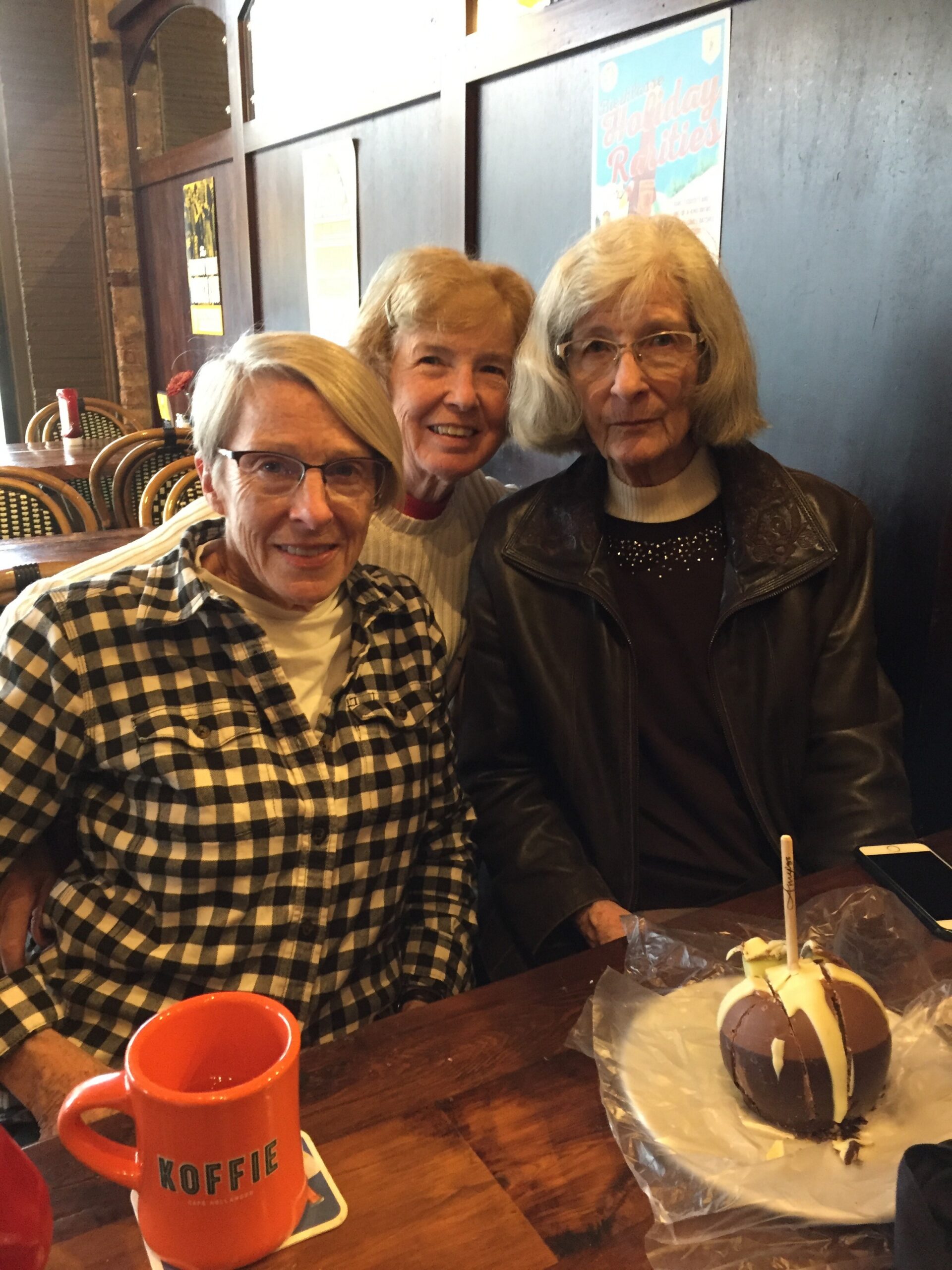 Three older women sit at a cafe table smiling at the camera, with a chocolate donut on foil and an orange mug labeled 'KOFFIE' in the foreground.