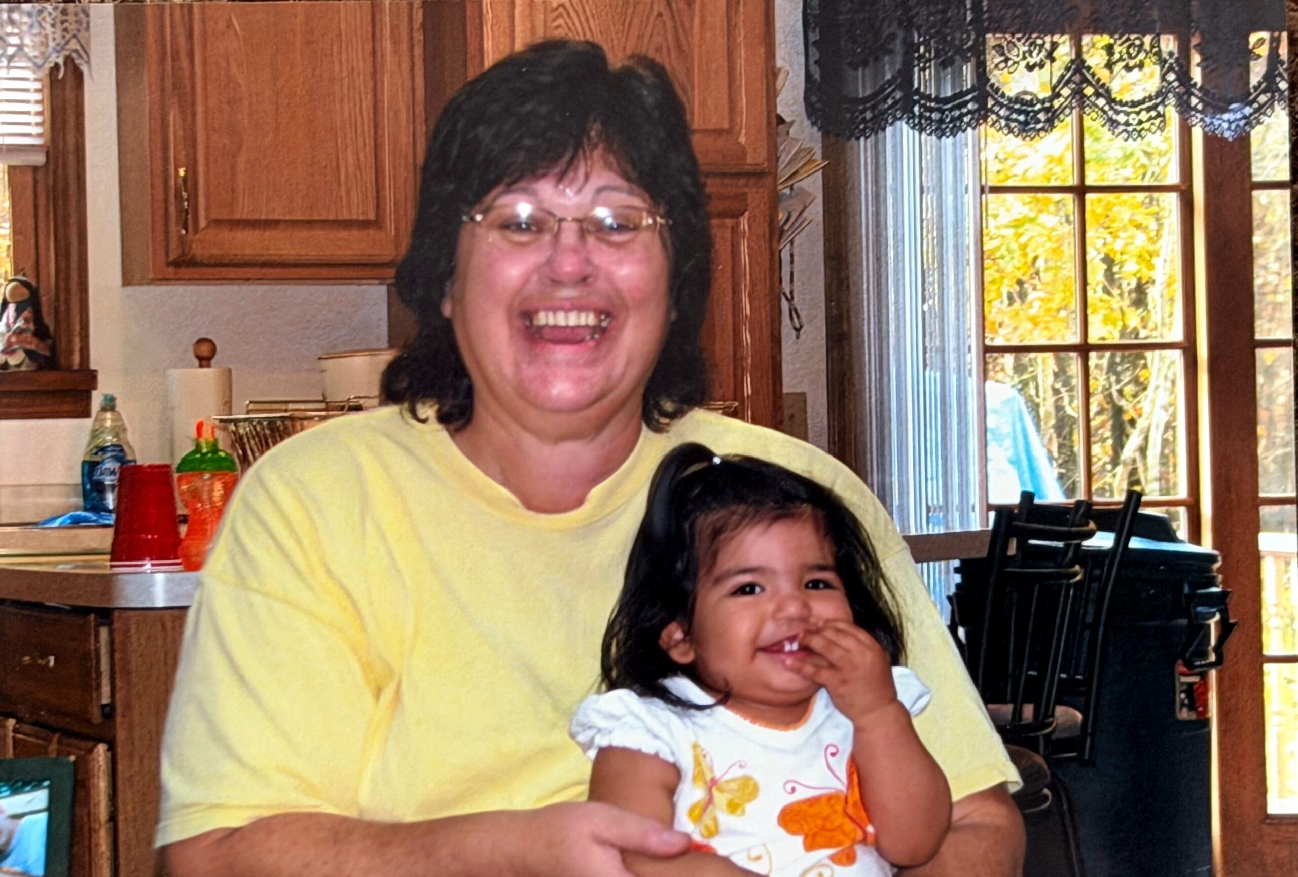 Smiling woman in a yellow shirt holding a laughing young girl in a kitchen with wooden cabinets in the background.