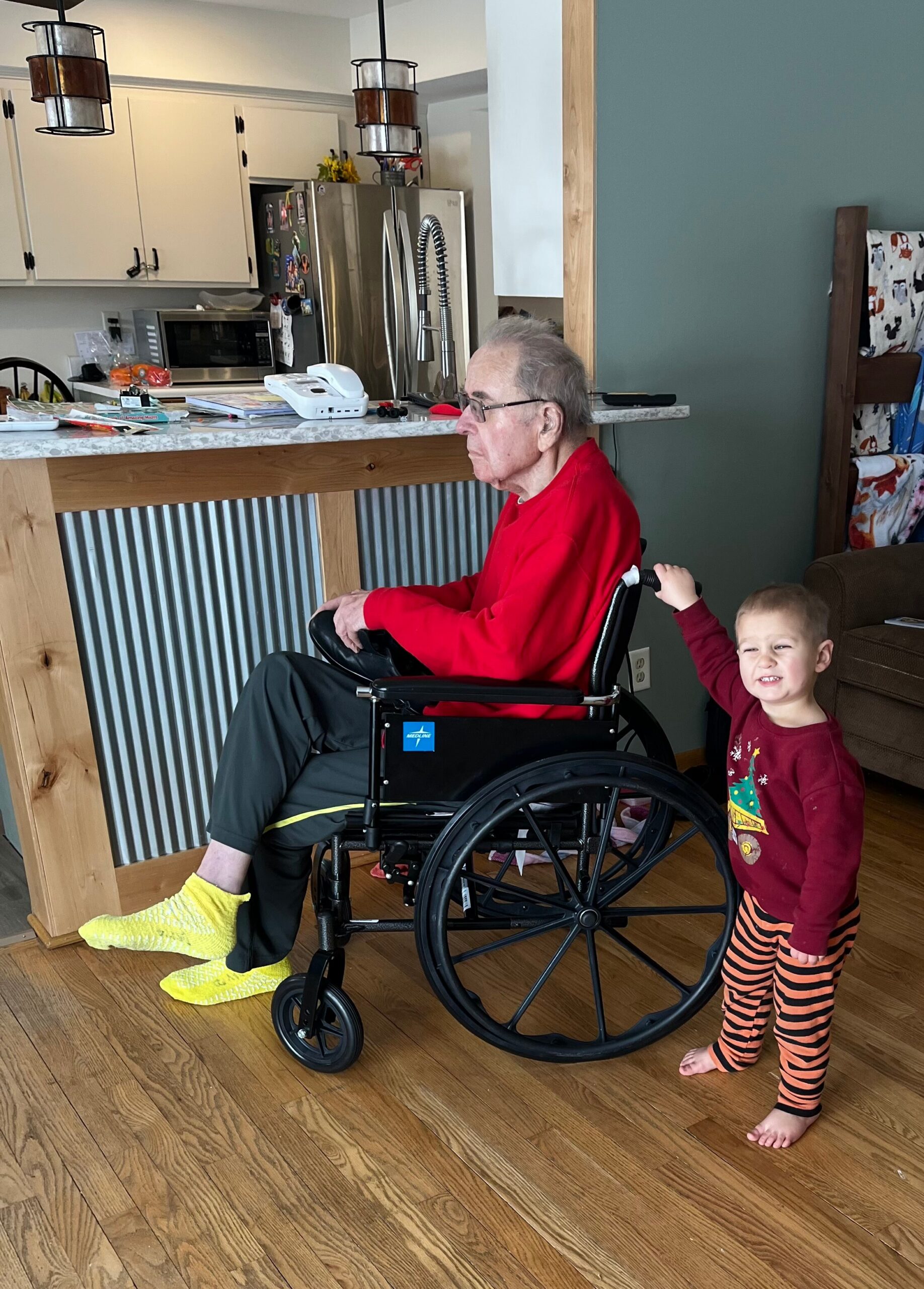 Elderly man in a red shirt sitting in a wheelchair at a kitchen island, with a young child standing beside him and holding the chair from behind.