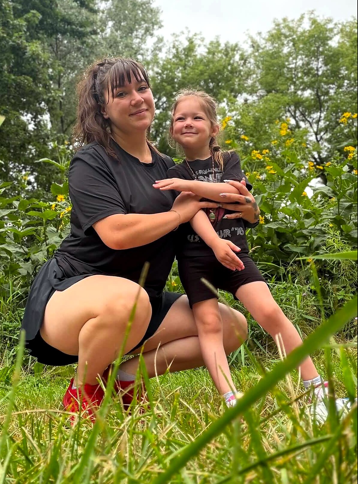 Woman kneeling in tall grass, holding a smiling young girl in a black outfit among yellow wildflowers in a sunny field.