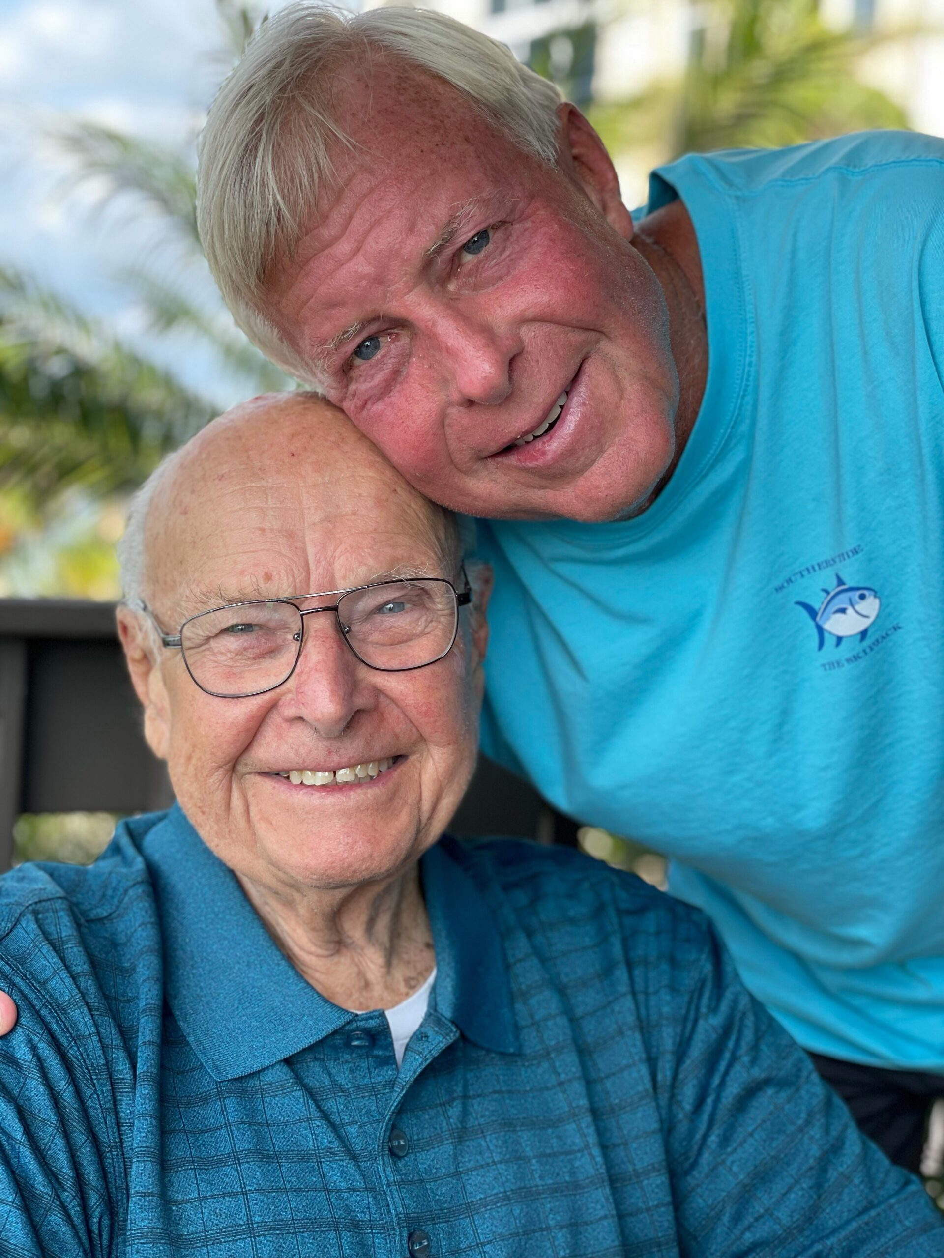 Two smiling older men posing closely together outdoors, one resting his head on the other’s shoulder.