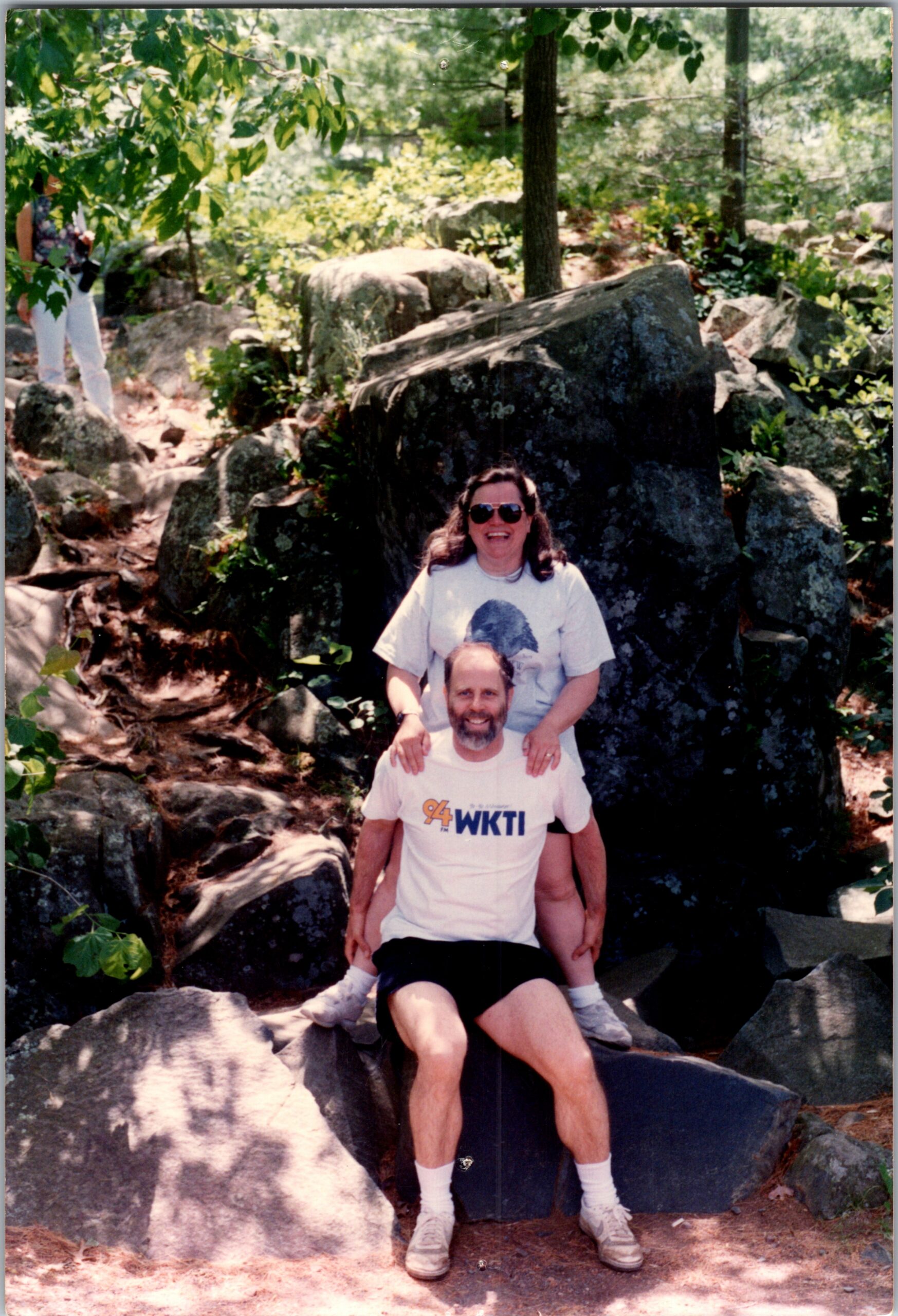 Two adults posing on large rocks in a sunny, wooded garden; a bearded man in a white WKTI T-shirt sits in front while a smiling woman wearing sunglasses stands behind him.