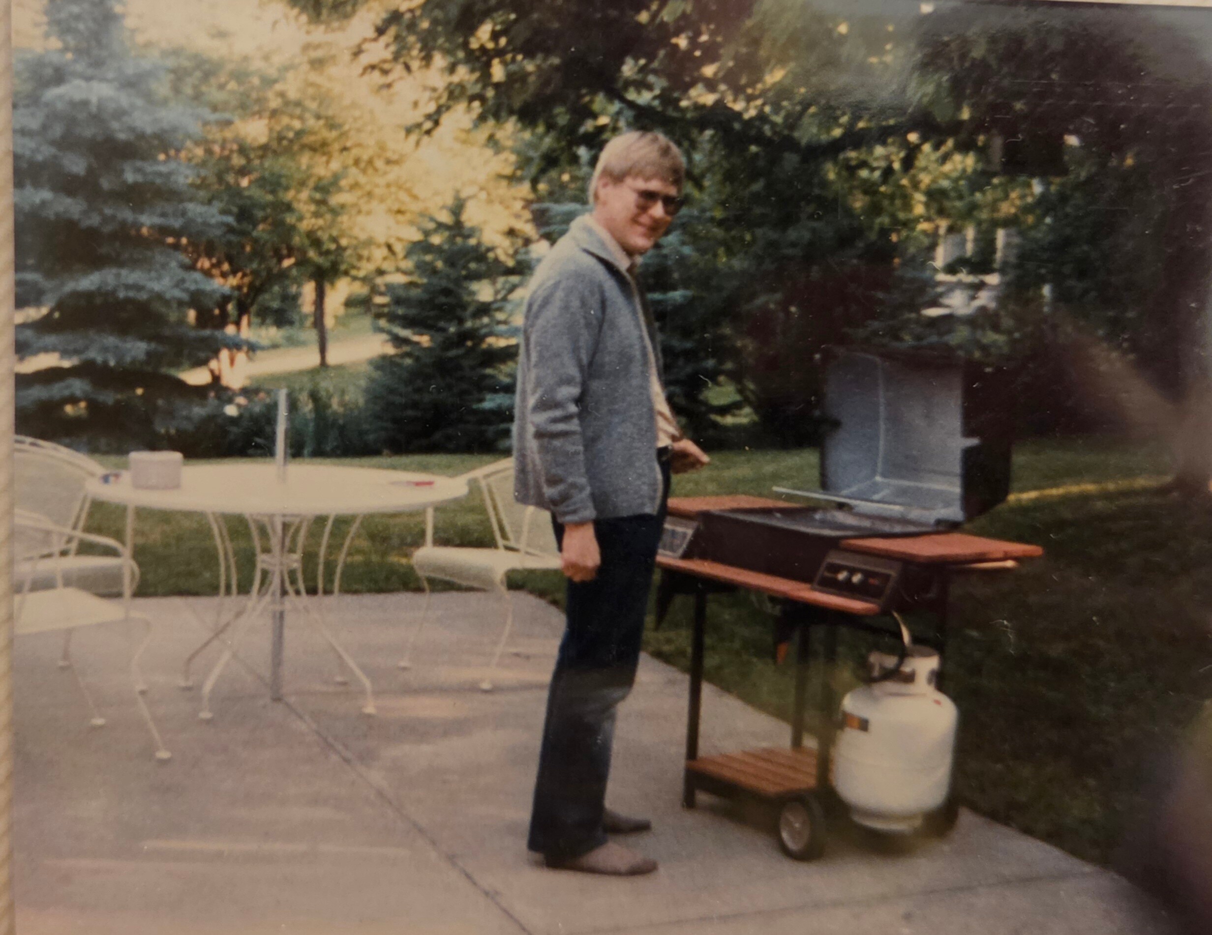 Man in a light jacket and sunglasses stands beside a barbecue grill on a tiled patio, with trees in the background.