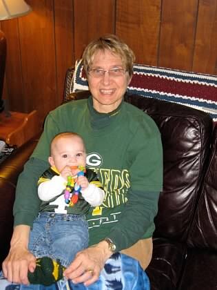 Older woman in a green sweatshirt sits on a leather couch with a baby on her lap, the baby holding a colorful toy.
