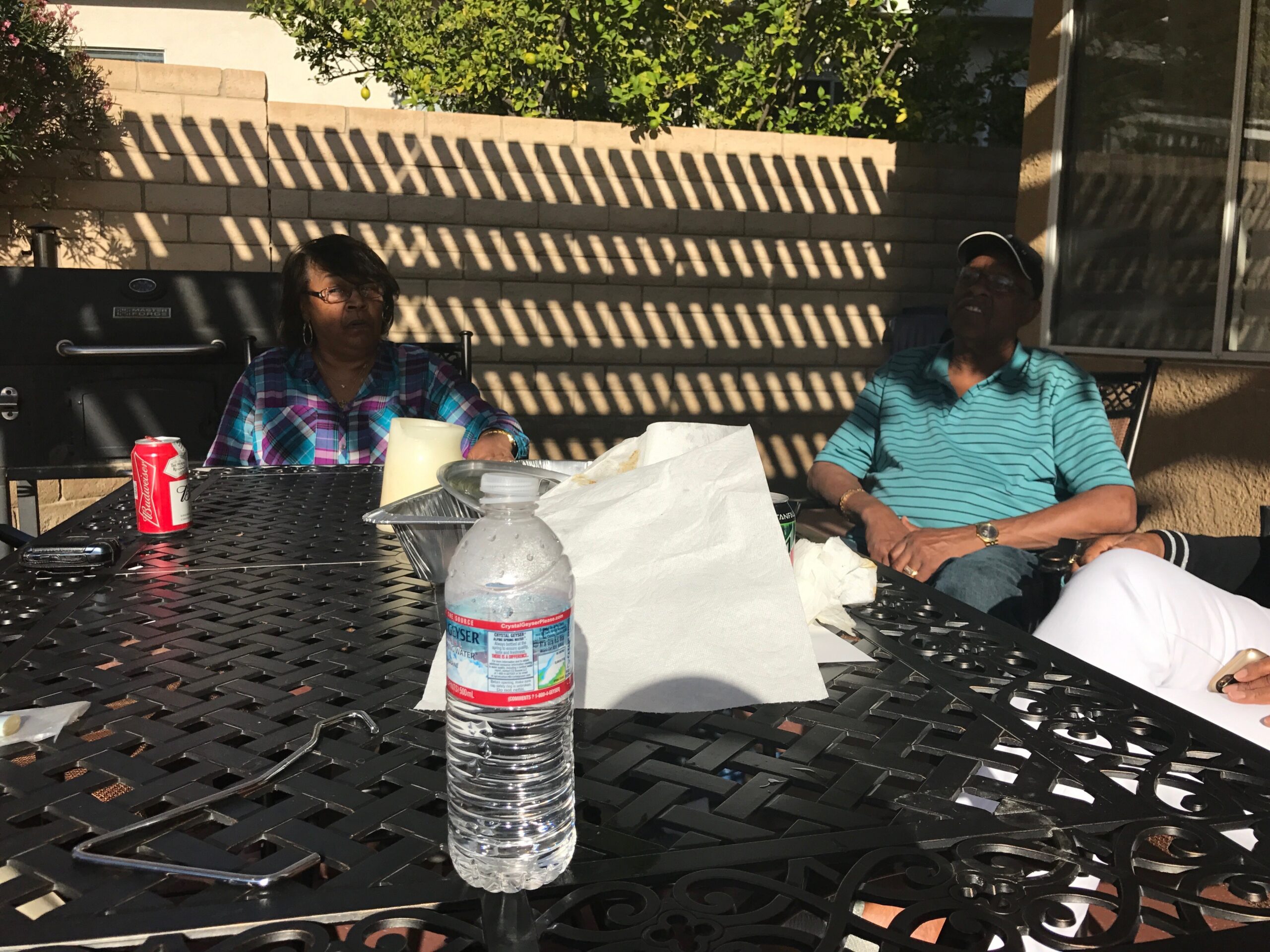Two people sit at a black metal patio table outdoors, with a brick wall and striped sun shadows in a backyard garden backdrop.