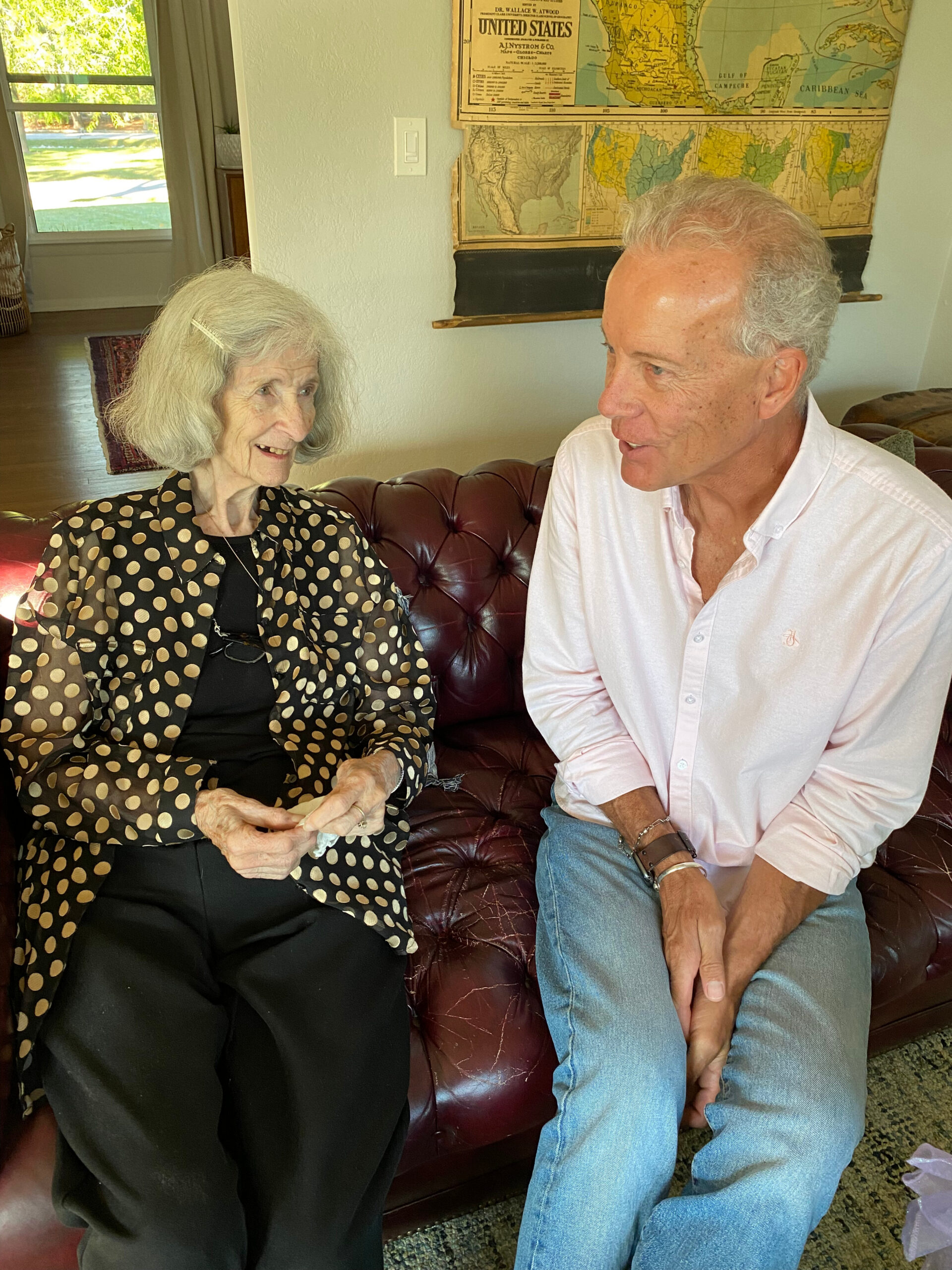 Two elderly people sit on a burgundy leather sofa, facing and talking in a sunlit living room with maps on the wall behind them.