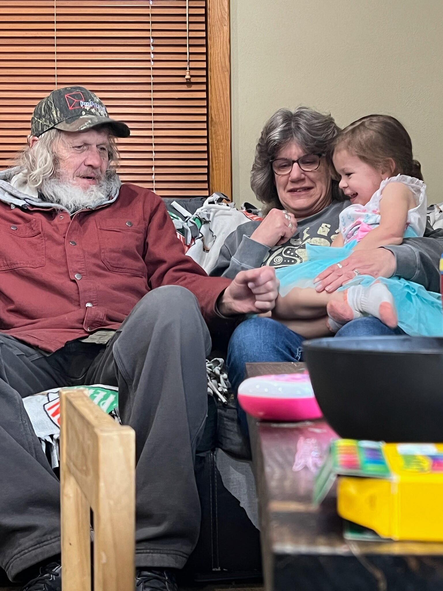Older man with a gray beard wearing a camo cap sits on a couch next to a smiling woman holding a young girl indoors beside wooden blinds.