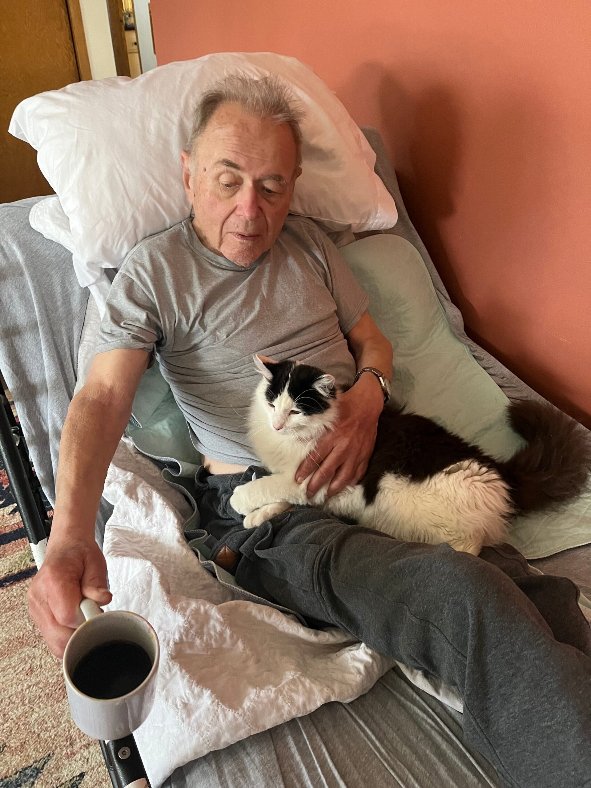 Older man sitting in a recliner with a black-and-white cat on his lap, a mug of coffee on a nearby tray.