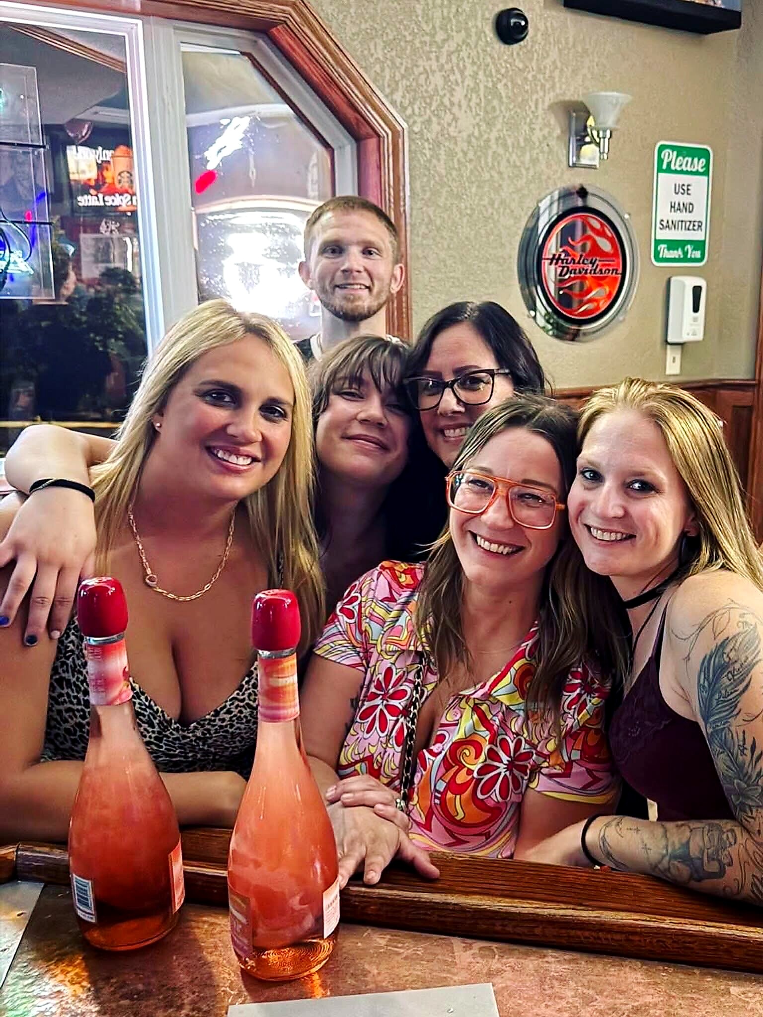 Group of friends at a bar taking a selfie, with two pink bottles on the counter.