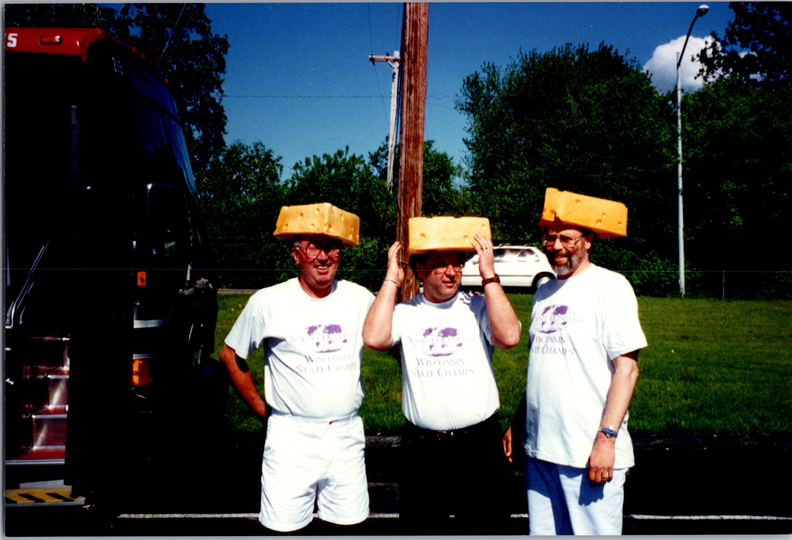 Three men wearing large yellow cheese blocks on their heads, posing outdoors at a sunny event, shirts read Wisconsin State Champs.