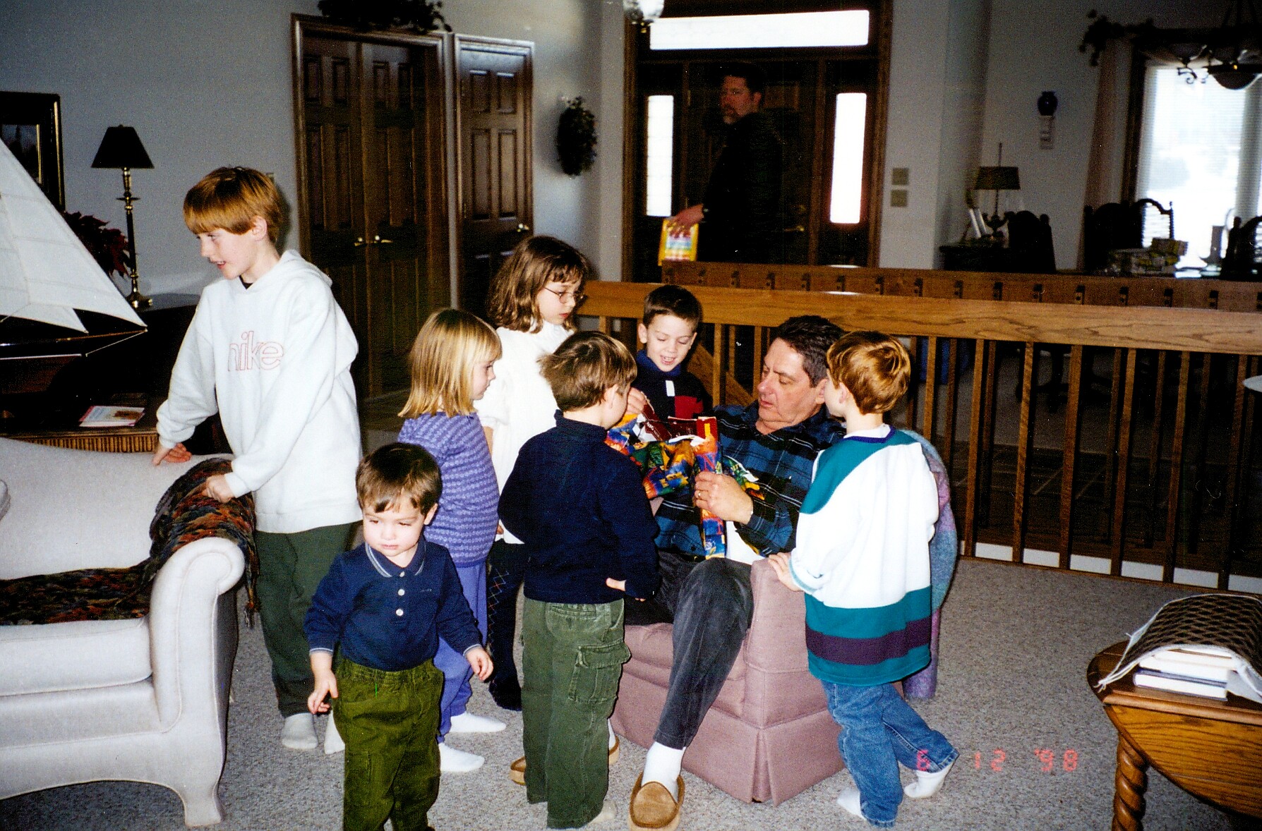Man sitting on a pink sofa hands out wrapped gifts to several children in a living room or family room setting.