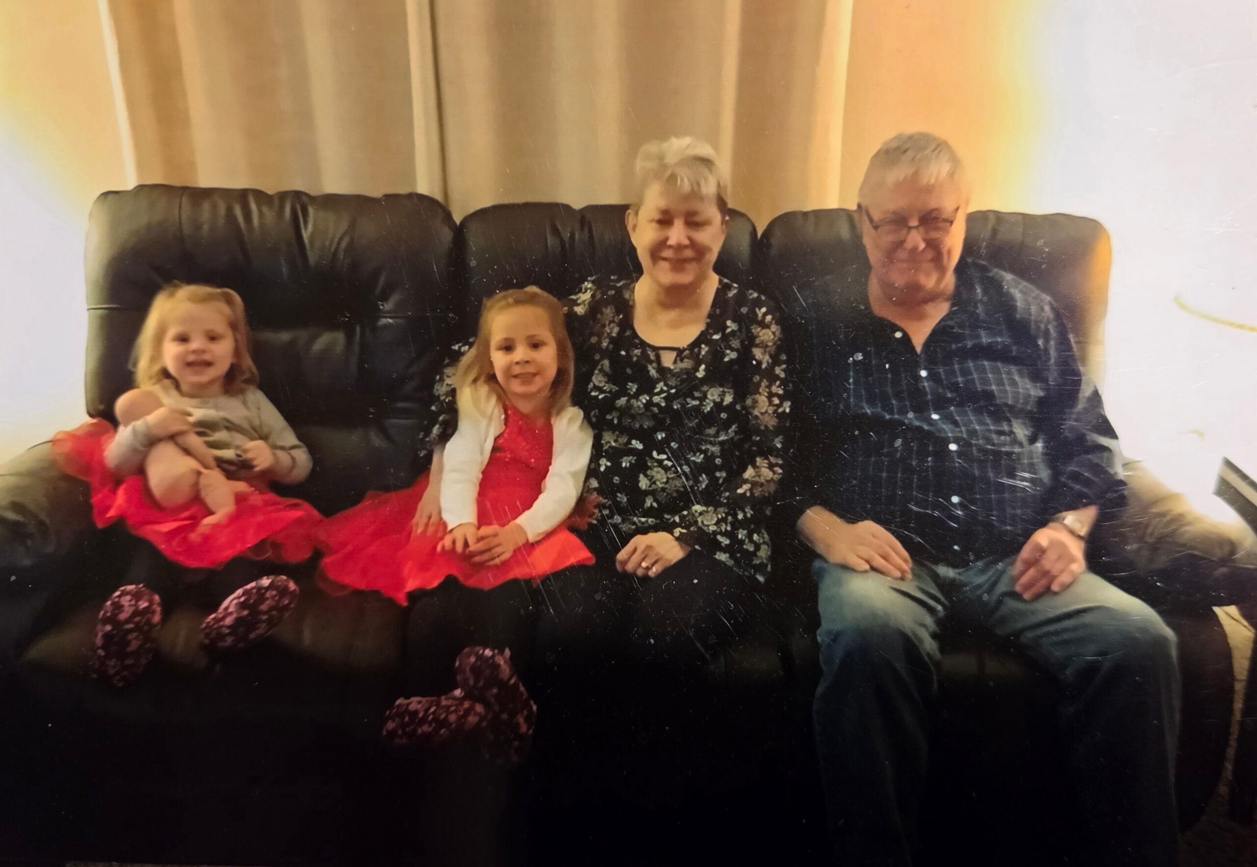 A family of four sits together on a black leather couch: two young girls in red dresses with smiles, flanked by an older woman and an older man.