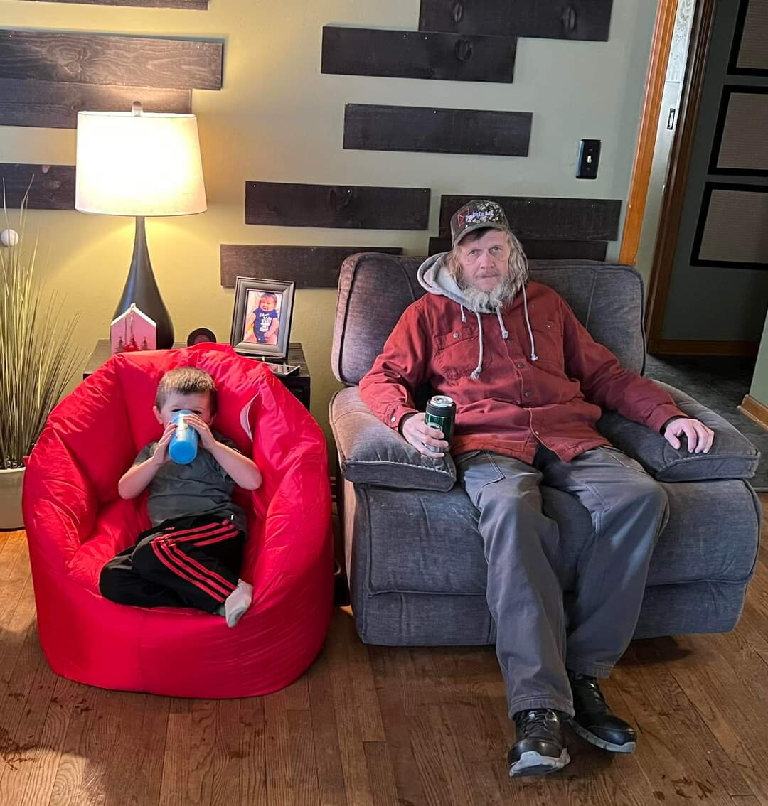 Man with a beard in a red jacket sits in a gray armchair while a child in a red bean bag drinks from a blue cup beside him, in a cozy living room.