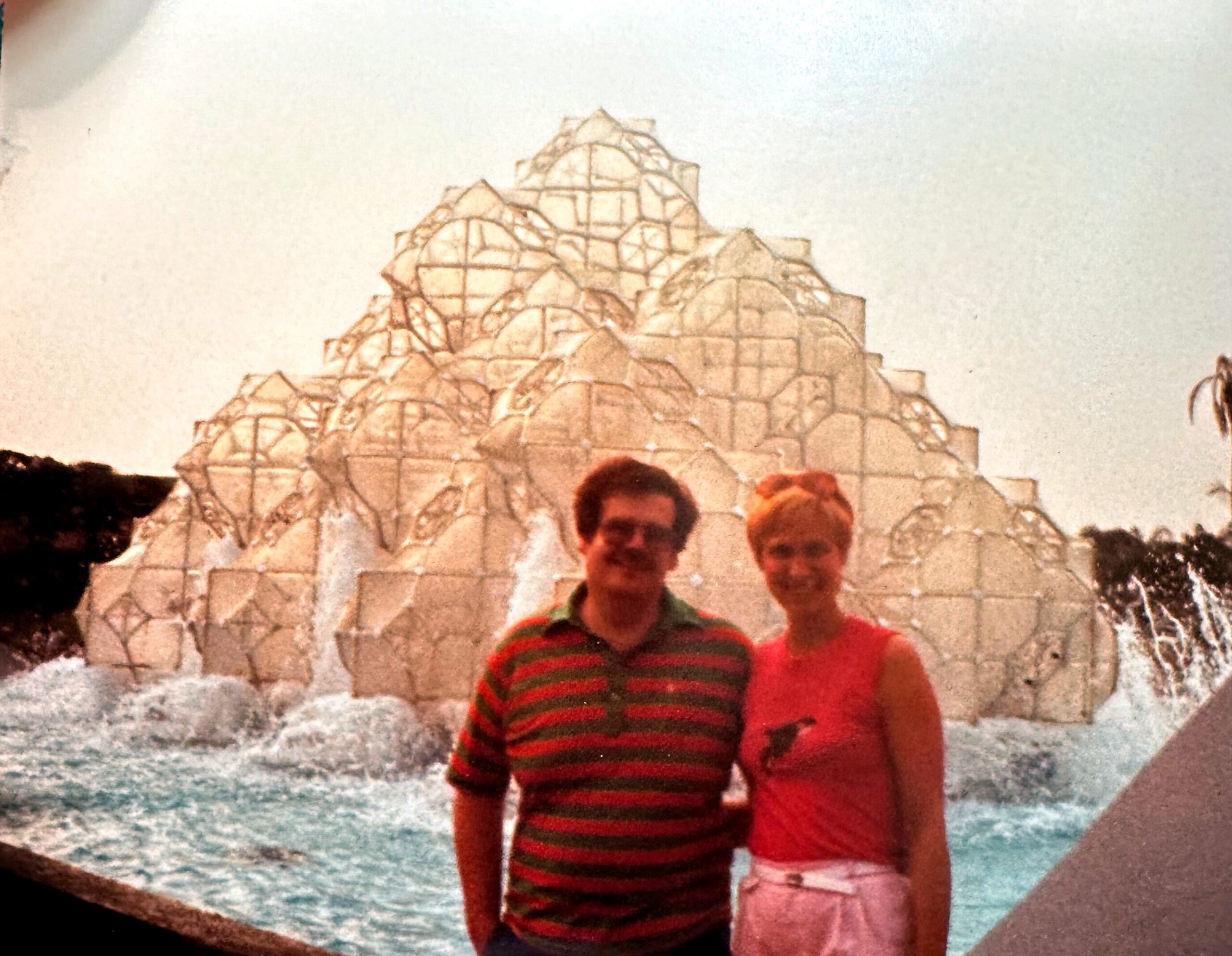 Two men posing together in front of a stepped stone pyramid sculpture with circular-arched details