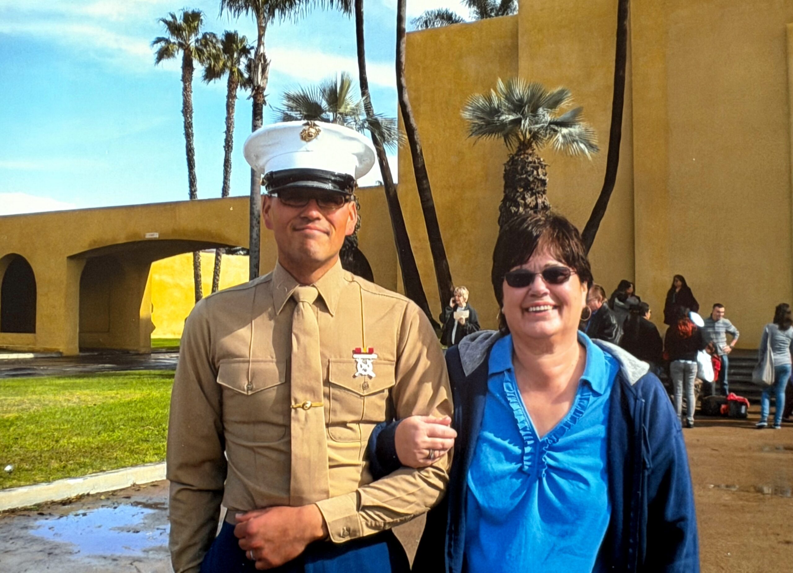 Man in a tan military dress uniform and white cap stands next to a smiling woman in a blue jacket, outdoors with palm trees and a yellow building in the background.