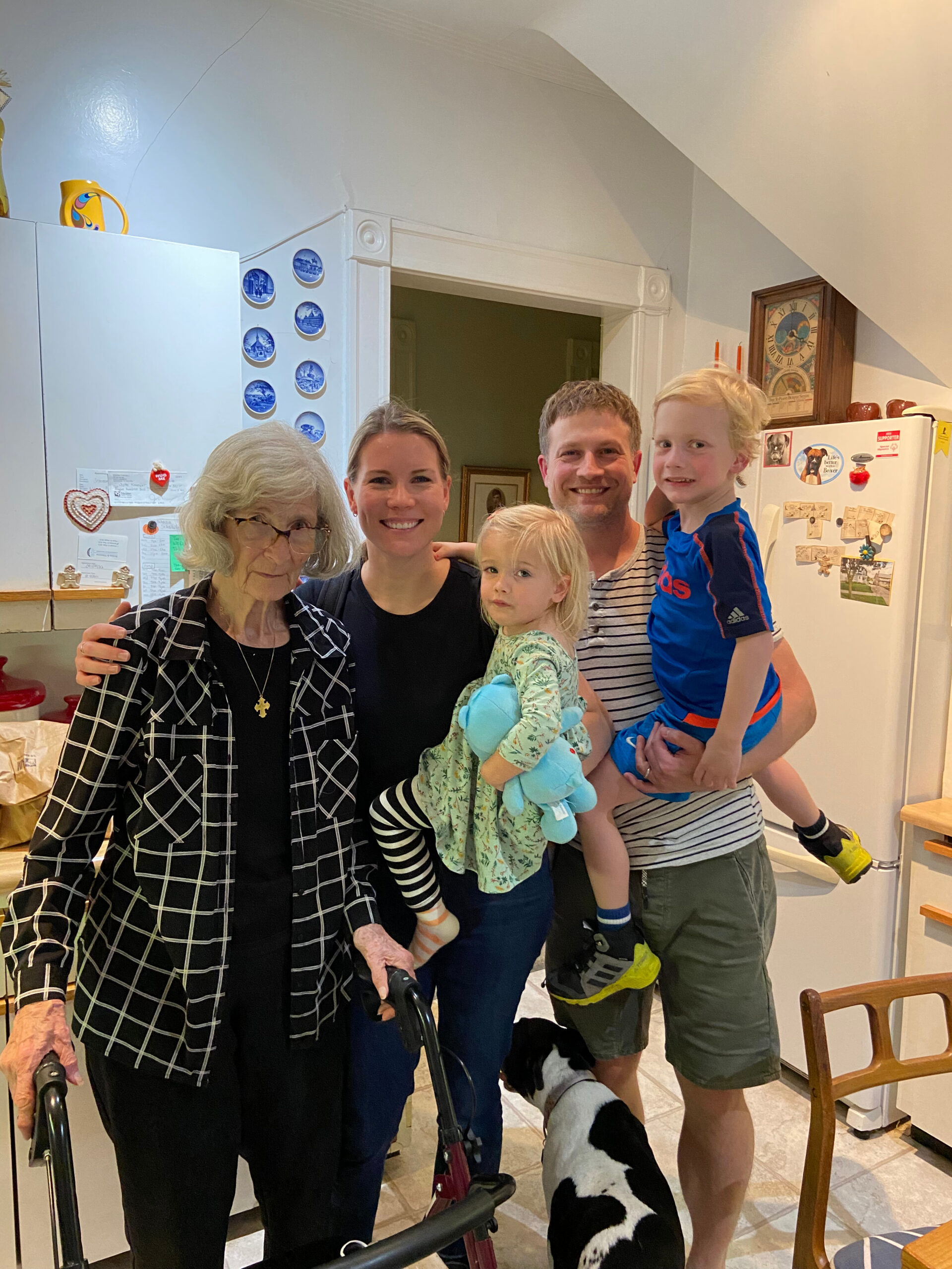 Family group photo in a bright kitchen: an elderly woman with a walker, two adults, two kids, and a black‑and‑white dog posing together.