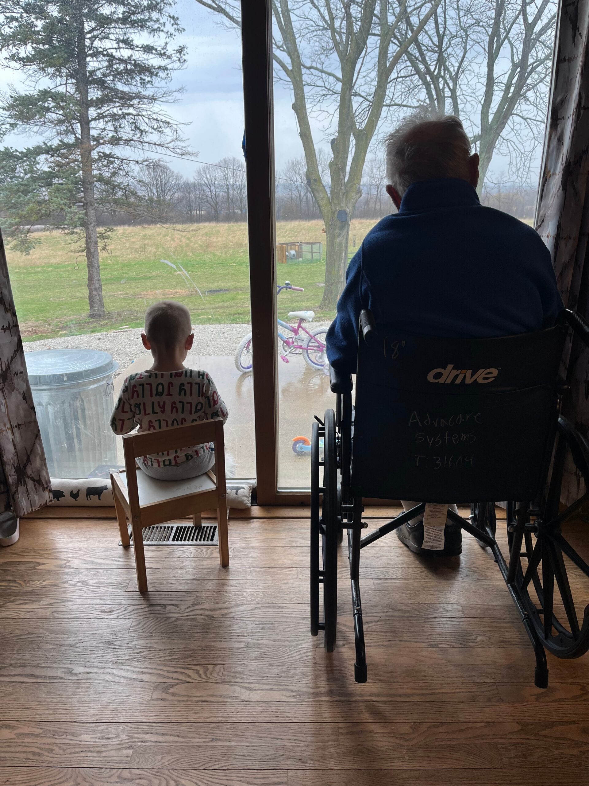 Child seated on a small wooden chair indoors beside a window, watching a yard with bare trees outside; a person in a blue wheelchair sits nearby.