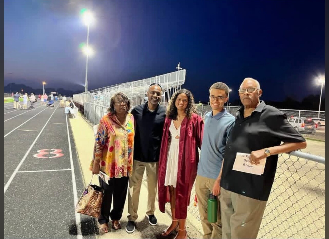 Group of five people posing on a track at night; one person in a maroon graduation gown and others in casual attire, smiling near a chain-link fence stadium bleachers in the background.