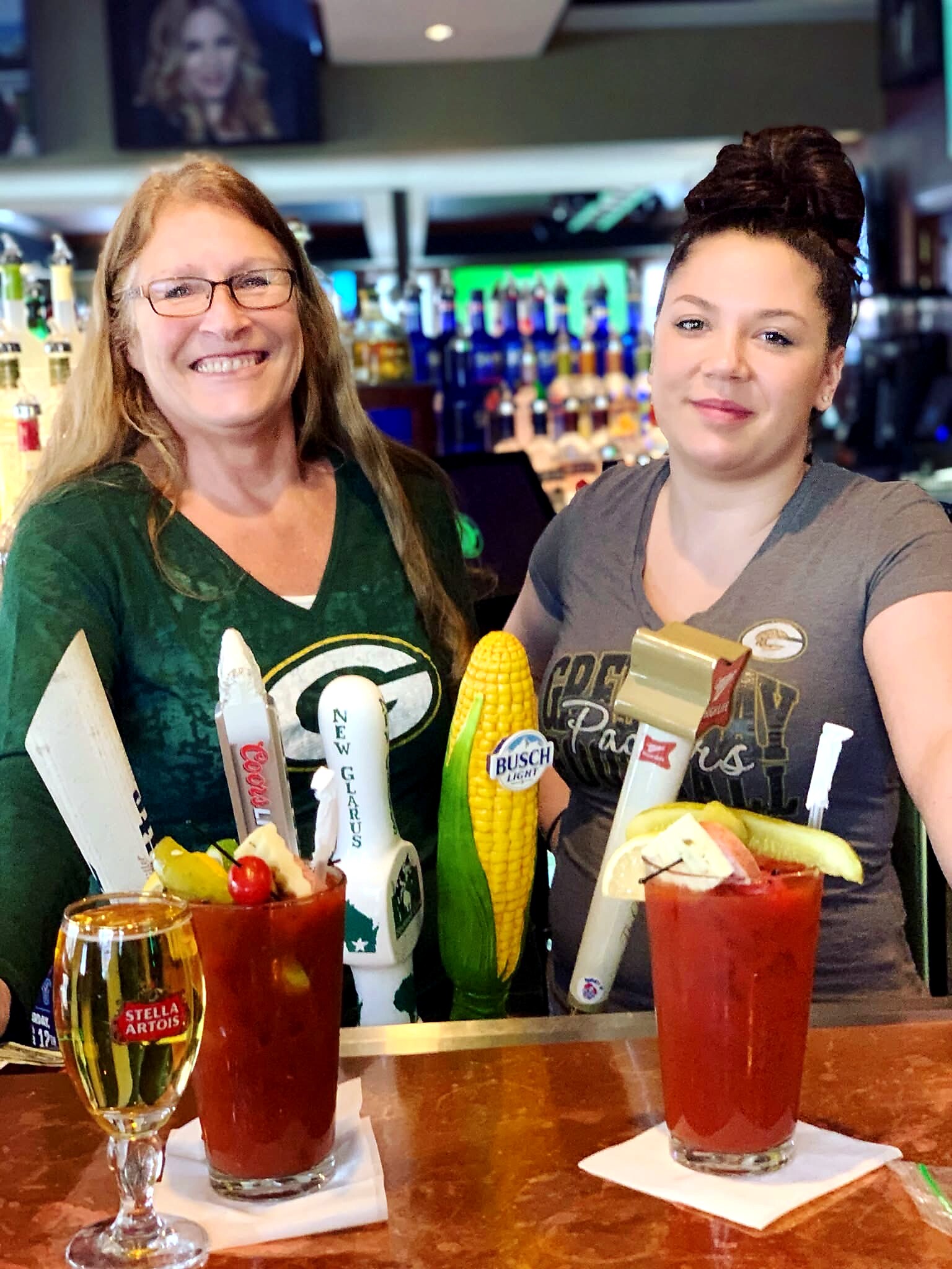 Two smiling women behind a bar with colorful drinks and corn cob garnishes, posing for a photo.”] ,