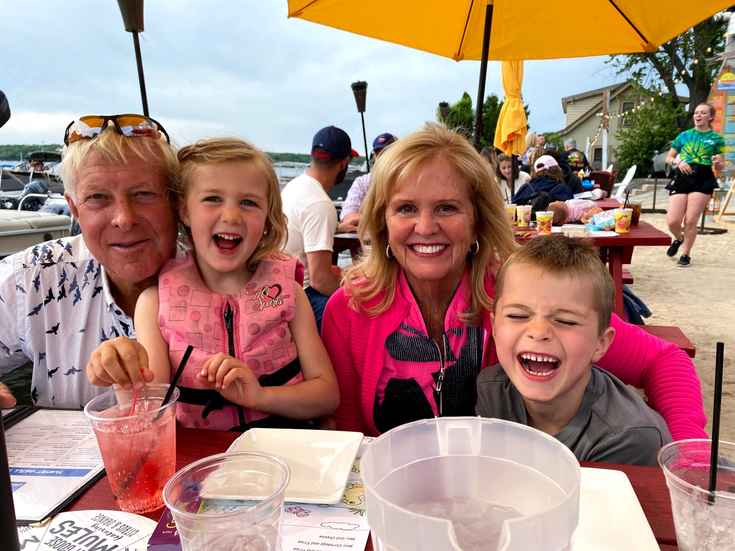 Family of four at a lakeside outdoor cafe under a large yellow umbrella, with pink drinks on the table and everyone smiling at the camera.