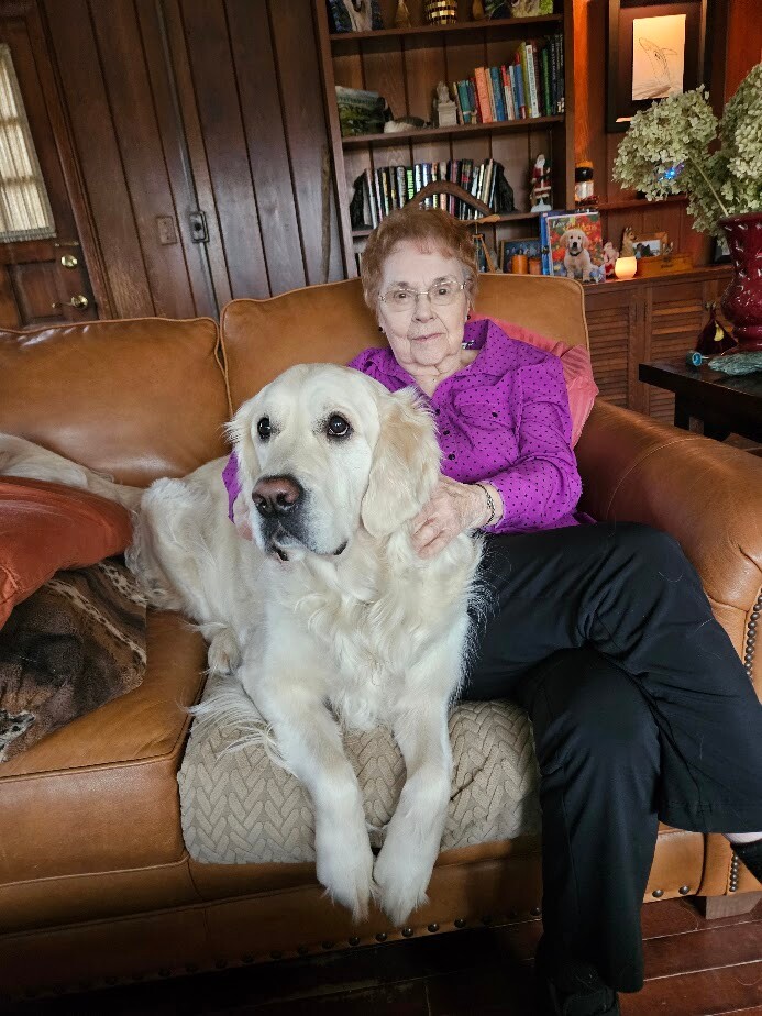 Elderly woman in a purple blouse sits on a tan leather sofa with a large white retriever resting its paws on the seat beside her.