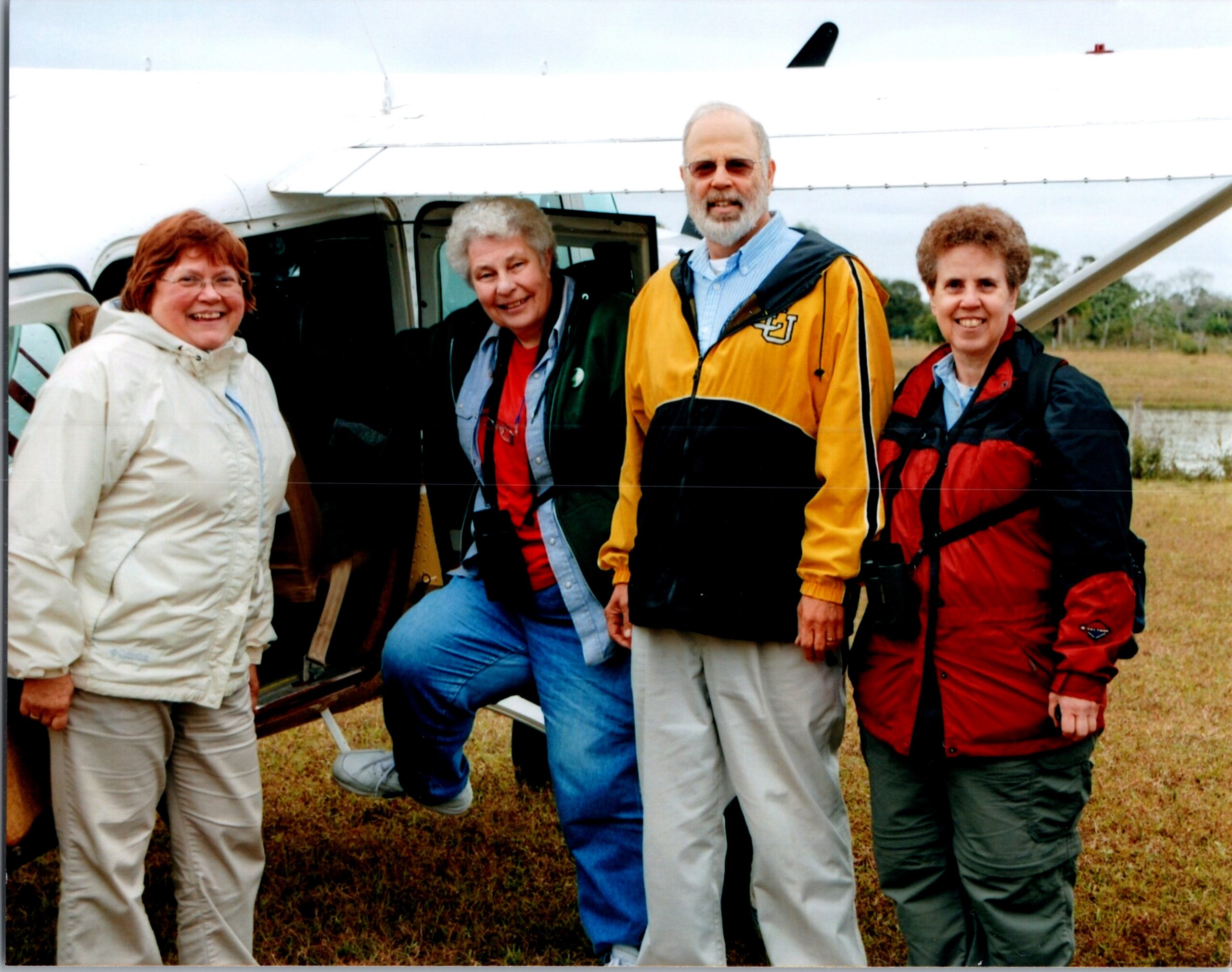 Group of four adults in outdoor jackets posing by a small airplane on a grassy field, smiling at the camera.