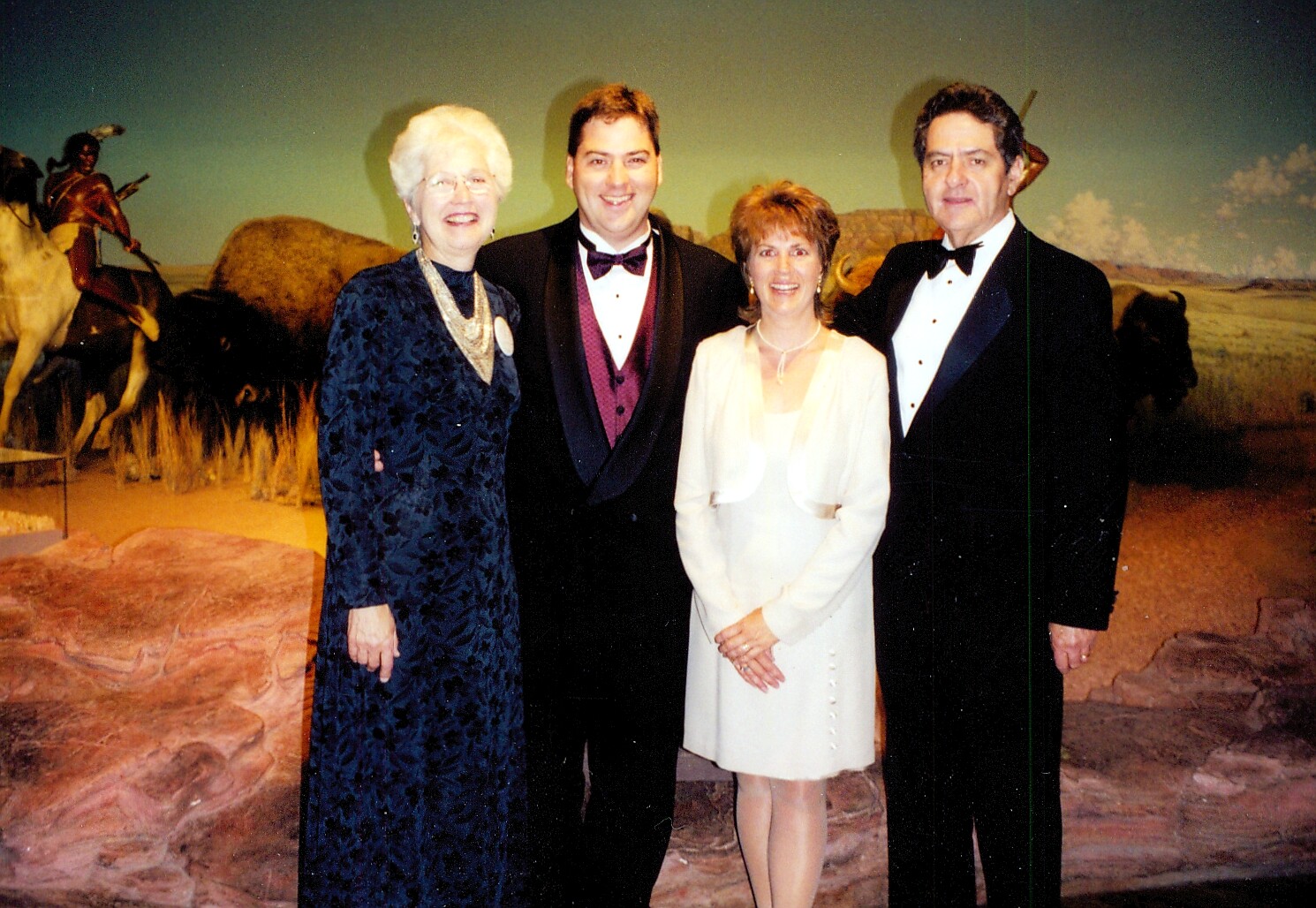 Four adults in formal evening wear pose together, smiling, in front of a desert mural with mounted horse figures behind them.