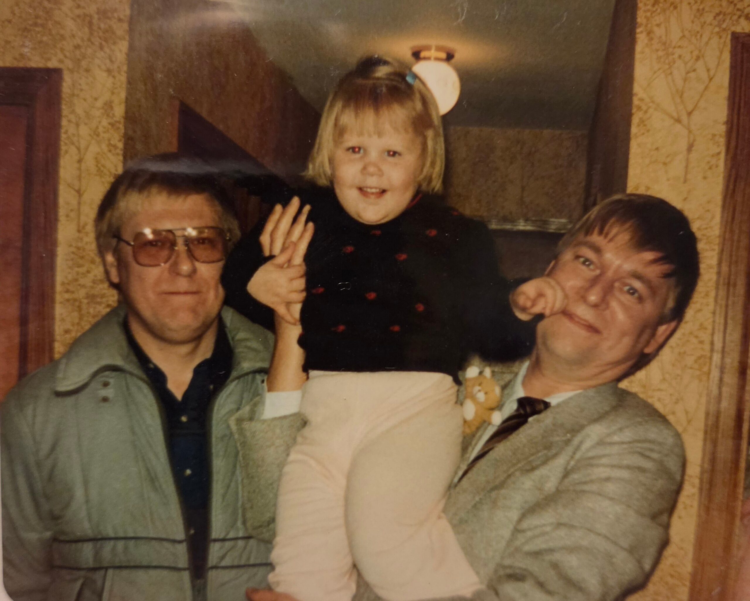 Young girl with blond hair sits on a man's shoulders, smiling, wearing a dark dress with small red accents, flanked by two adults in a home setting.