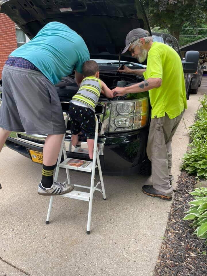 Man in neon shirt and cap works under the hood of a black SUV as a child on a step stool looks into the engine with another adult nearby.