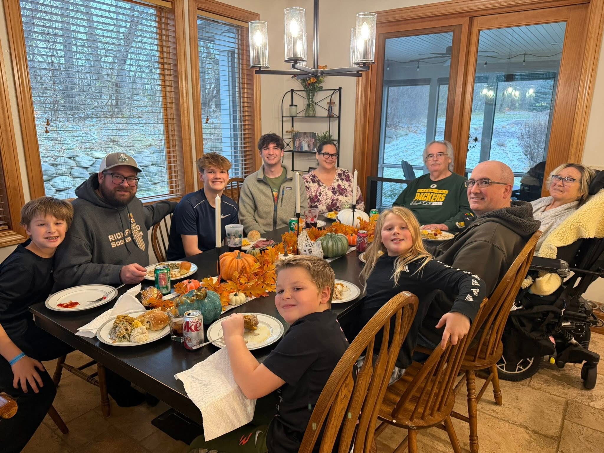 A large multi-generational family sits around a dining table with fall pumpkins and candles, eating indoors as autumn leaves show outside the windows.