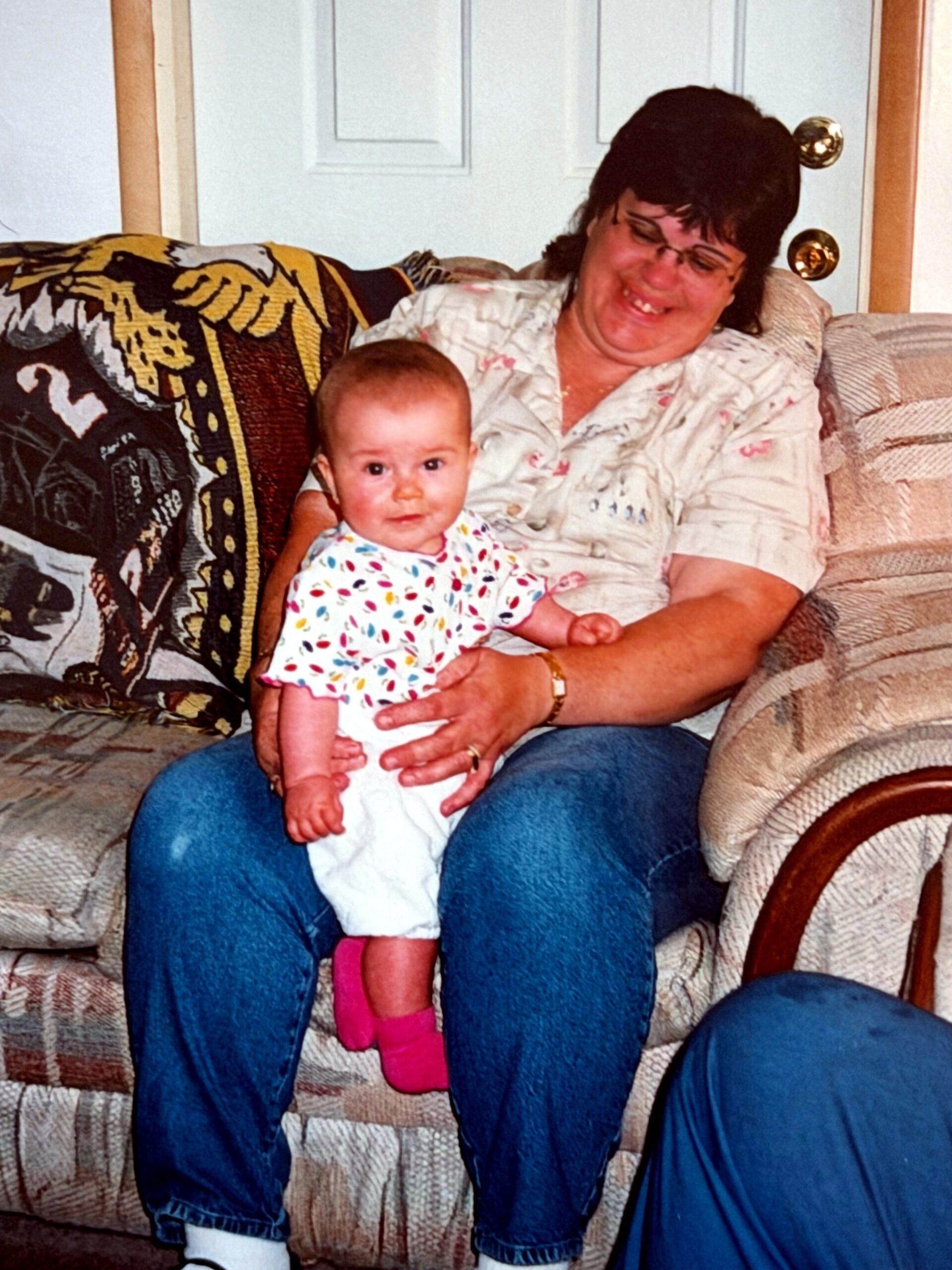 Grandmother sitting on a patterned sofa, smiling, while holding a baby girl in a white outfit with colorful prints.