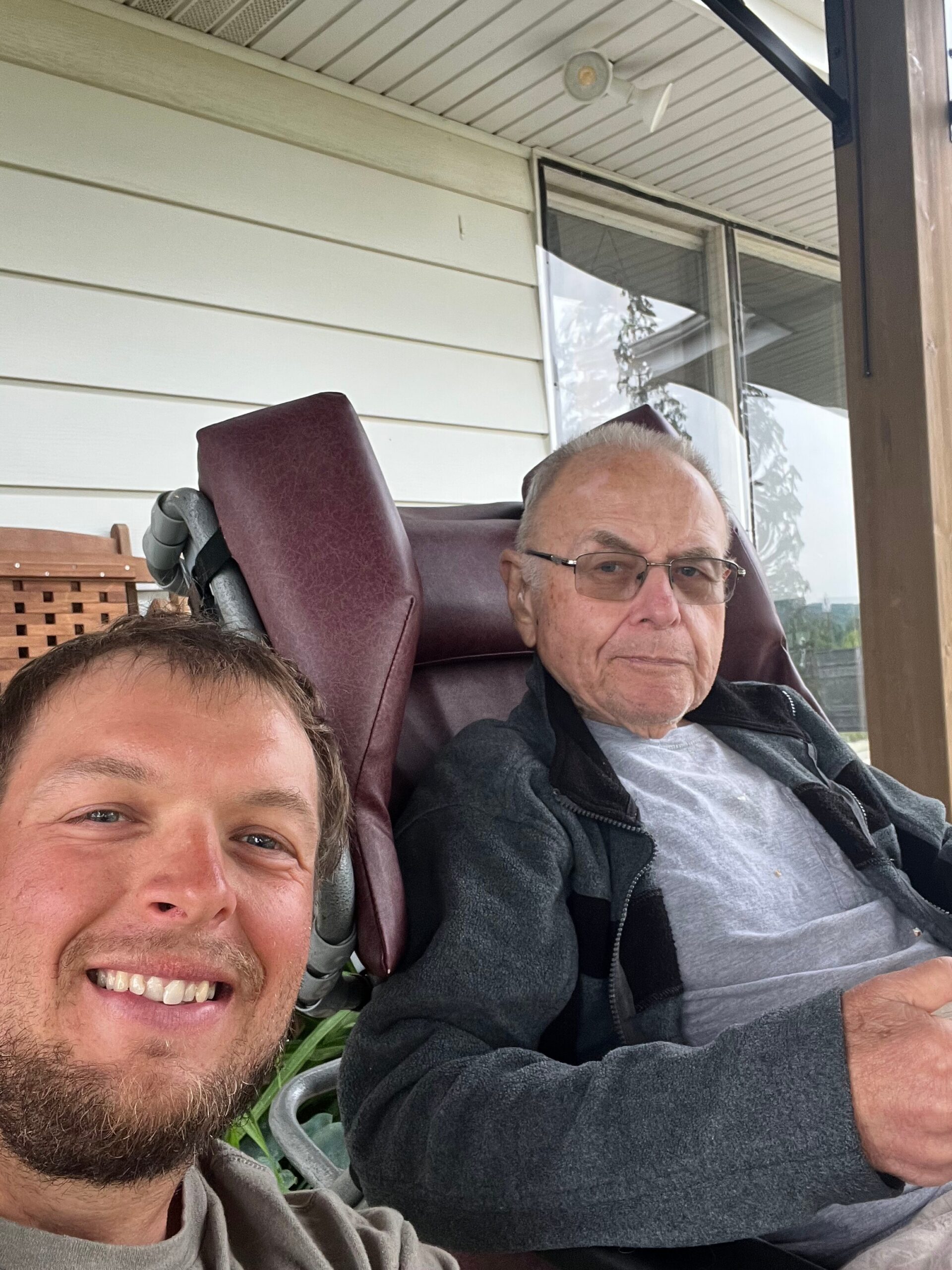 Two men posing for a selfie on a porch; younger man in front smiling, older man in a burgundy recliner wearing glasses in the background.