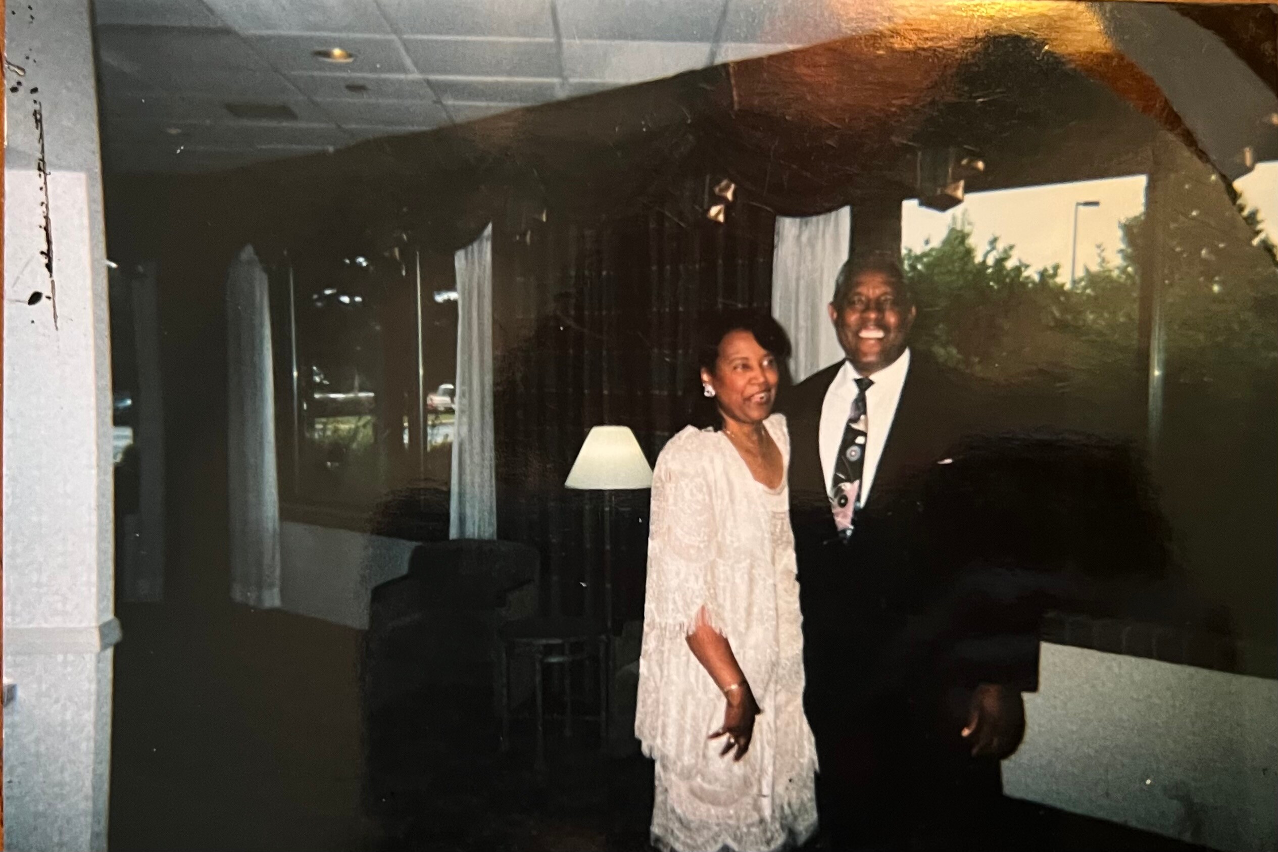 Smiling couple in formal attire posing indoors; woman in a light lace dress and man in a suit with a patterned tie, standing under an archway with a lamp in the background.
