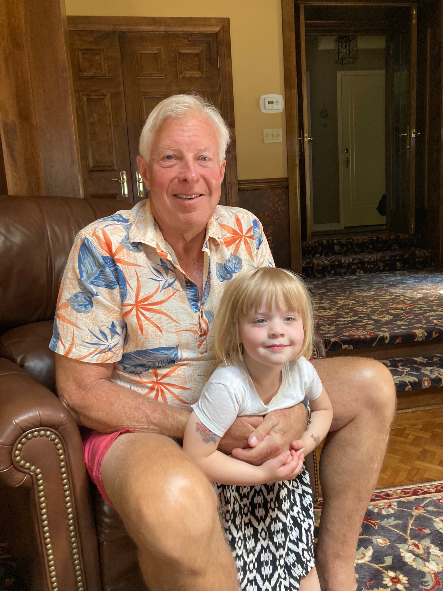 Older man with white hair smiles as he sits on a brown leather chair, a young girl on his lap, in a warmly wood-paneled room.