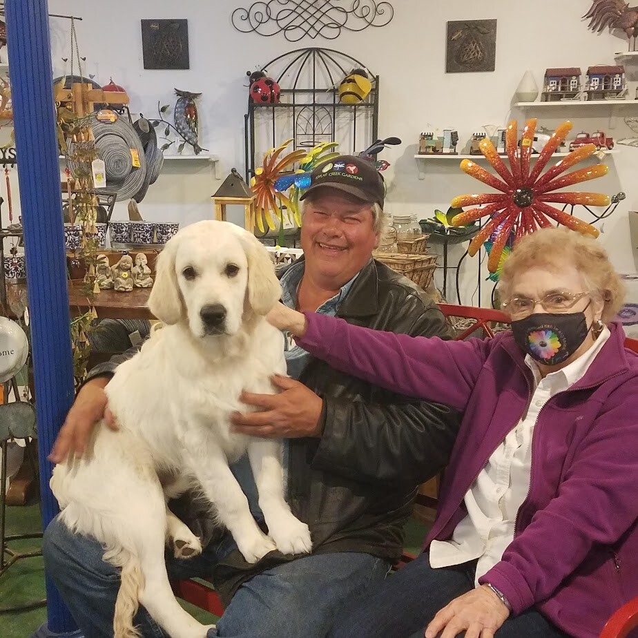 Smiling man and woman sit with a fluffy white dog in a colorful gift shop filled with ornaments and decorations on shelves behind them.