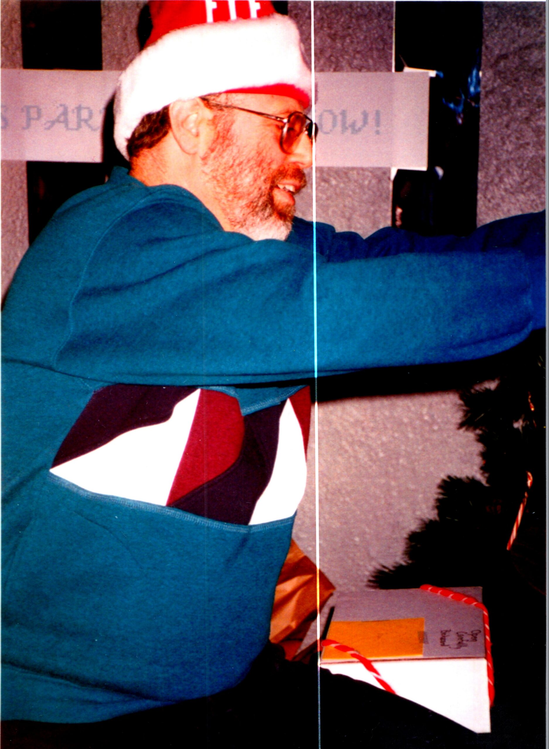 Man wearing a Santa hat and teal sweater, smiling, with wrapped gifts nearby.